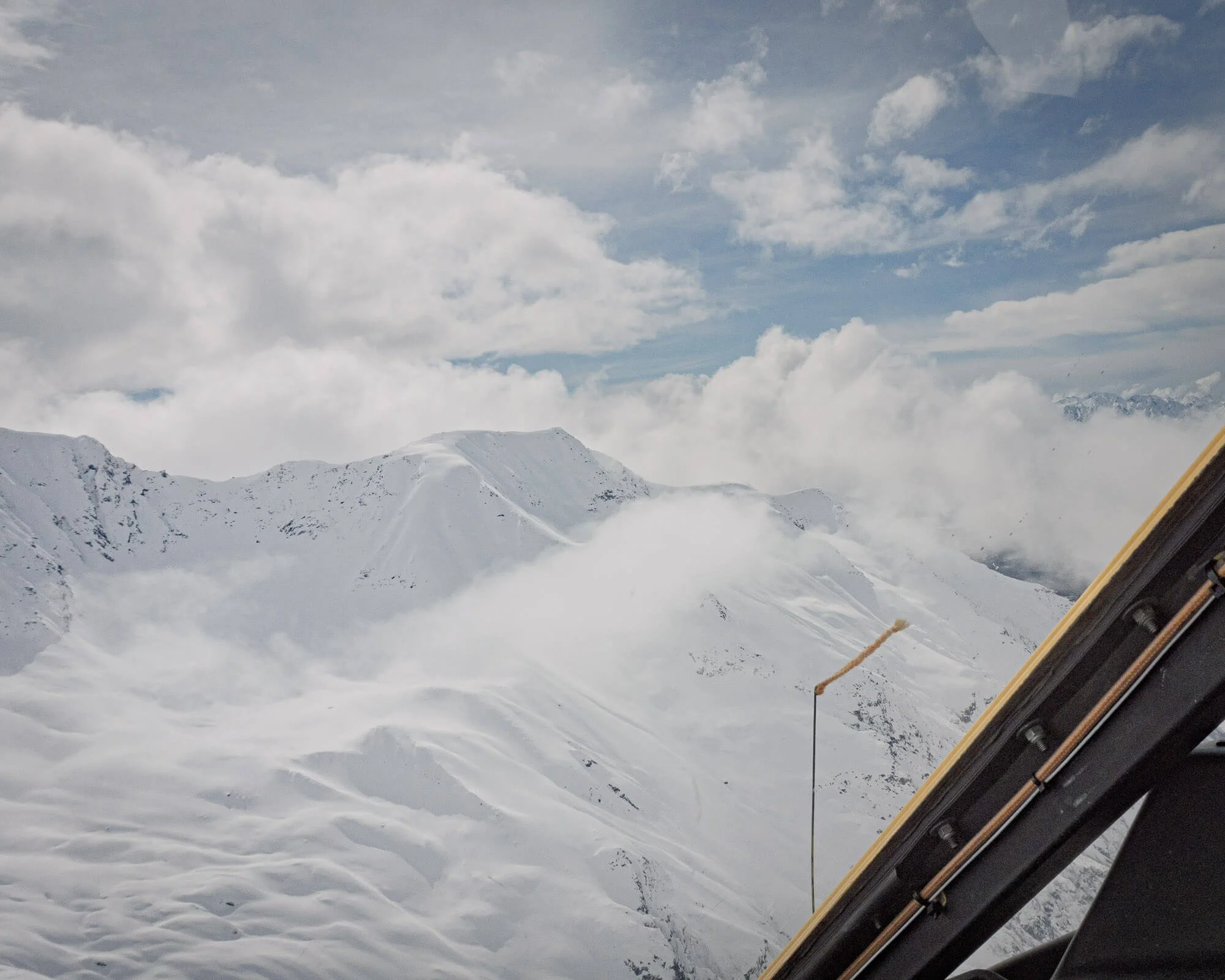 Snow-covered mountain range seen from an aircraft window with part of the aircraft's interior visible in the foreground.