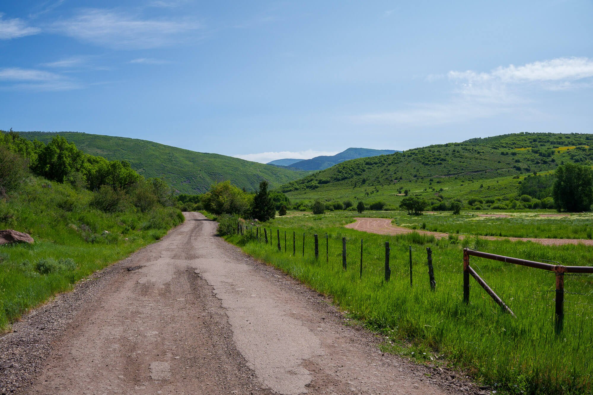 A dirt road winding through lush green countryside with rolling hills, trees, and a blue sky with scattered clouds.