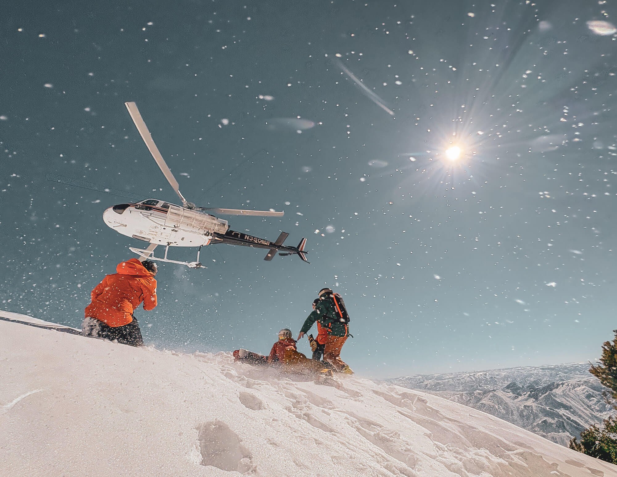 A helicopter flying in a snowy mountainous landscape with three people in winter gear on the snow, under a clear blue sky with the sun shining brightly.