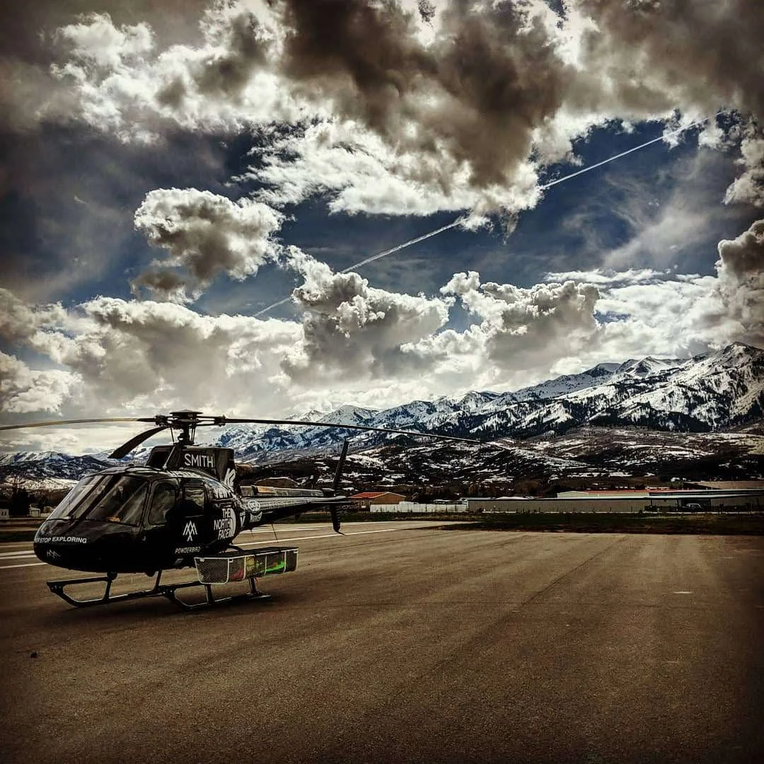 A black helicopter with markings and company logos, parked on an empty runway, with snow-capped mountains and a cloudy sky in the background.