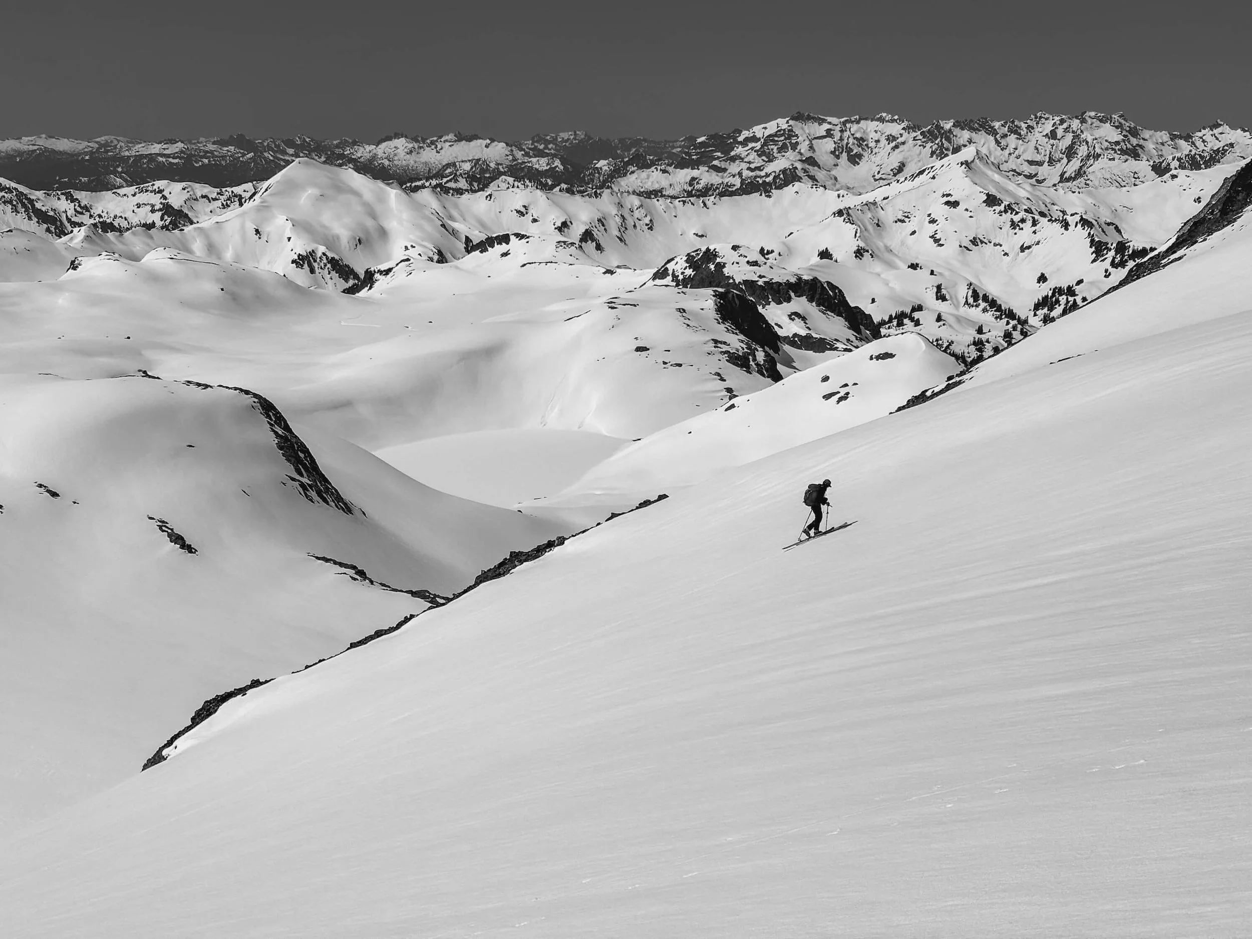 A lone skier descending a snowy mountain slope with a vast mountain range in the distance.