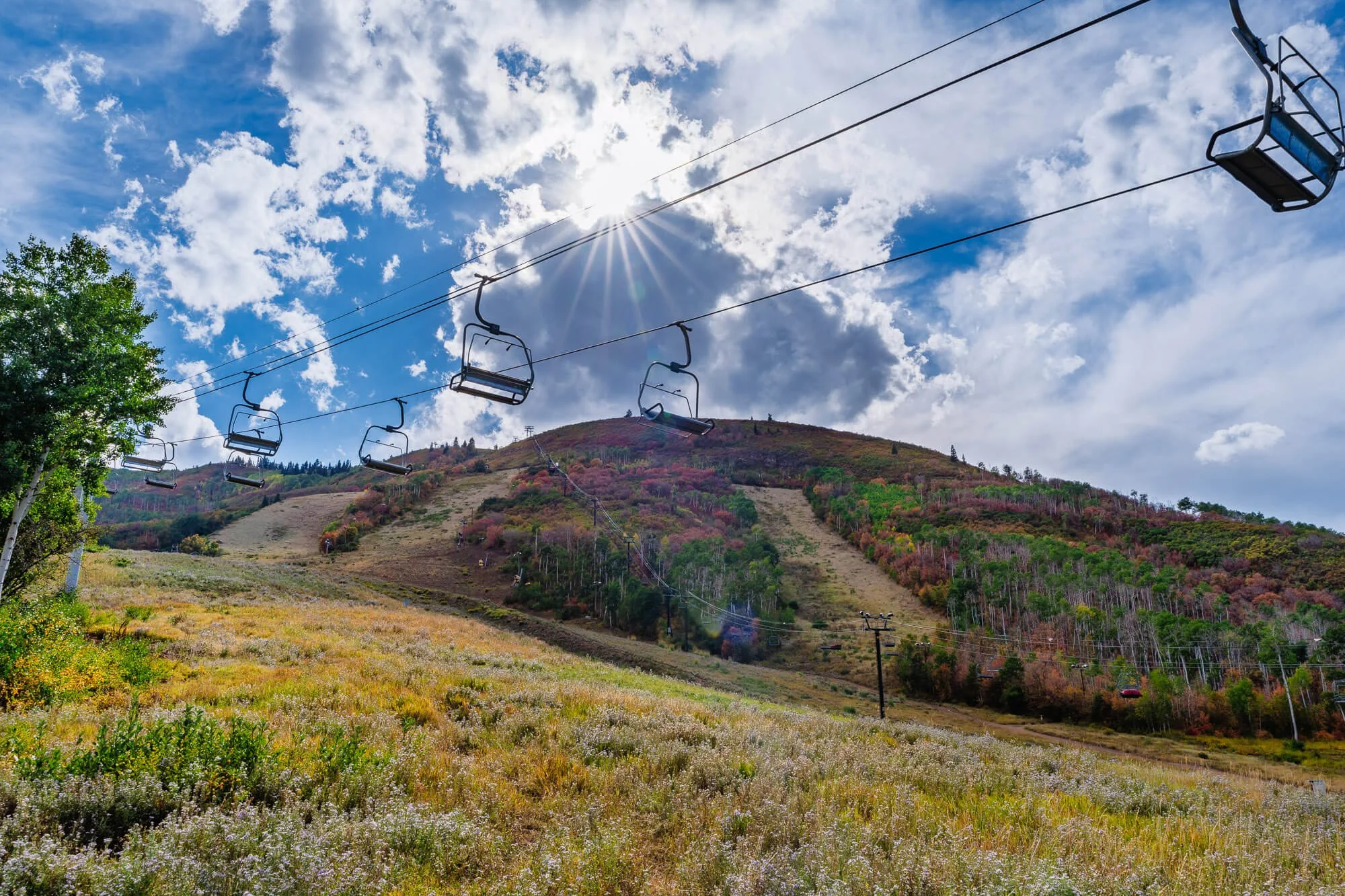 Ski lift chairs on a hill with colorful foliage and a partly cloudy sky.