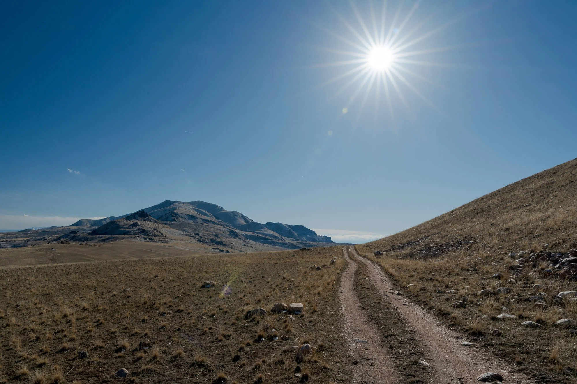 Dirt road leading toward distant mountain landscape under a bright sun in a clear blue sky.