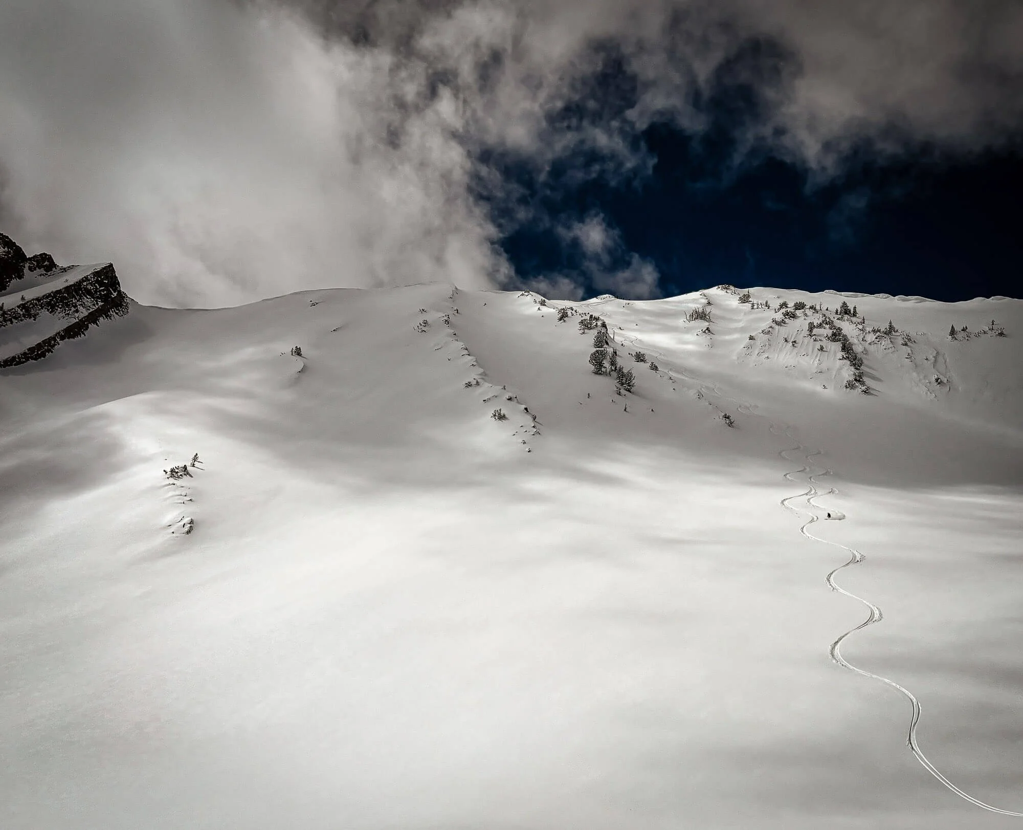 Snow-covered mountain slope with ski or snowboard tracks, small trees, clouds in dark sky.