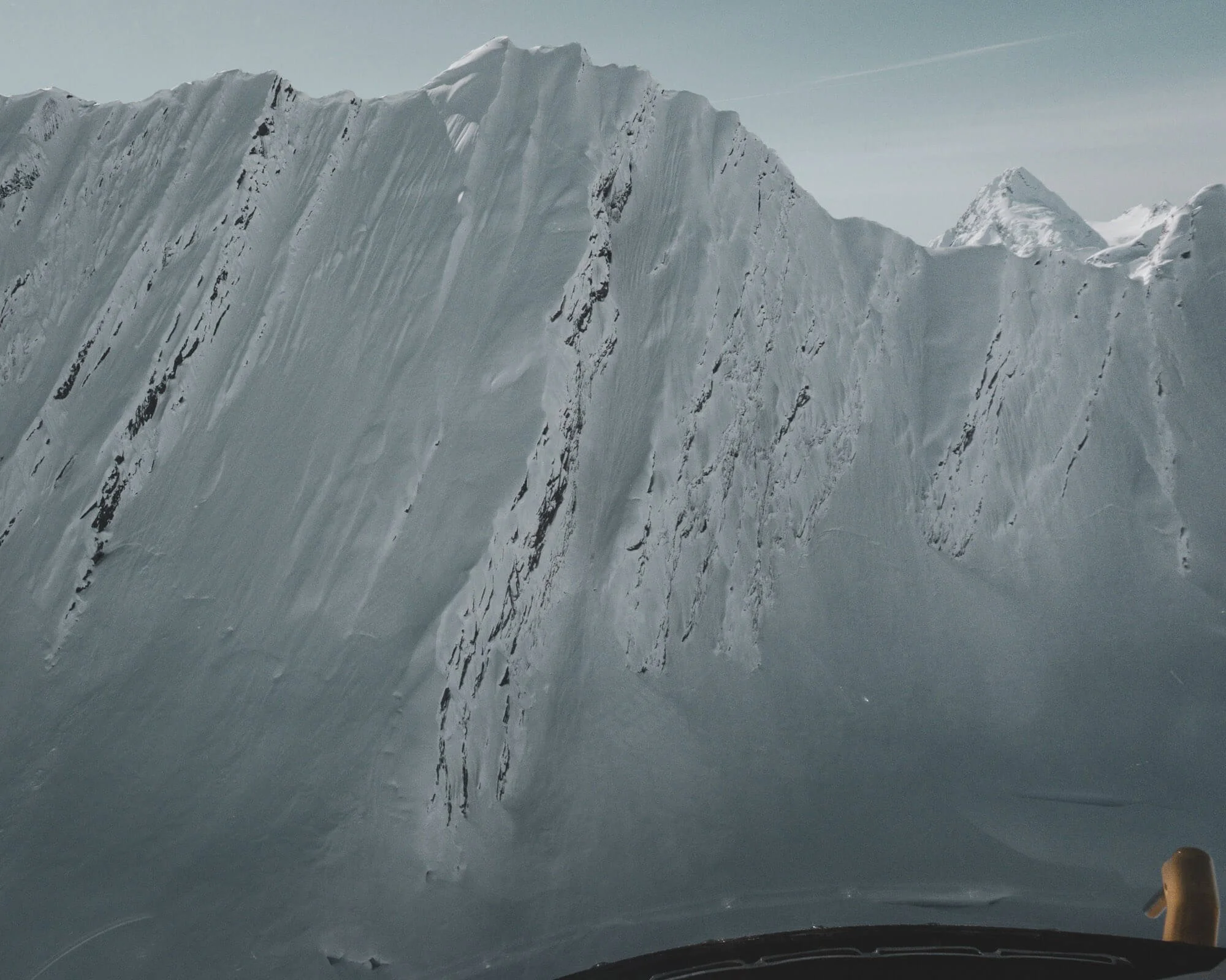 View of a large snow-covered mountain or glacier with a steep, jagged edge, taken from inside a vehicle or aircraft with part of the dashboard or cockpit visible at the bottom.