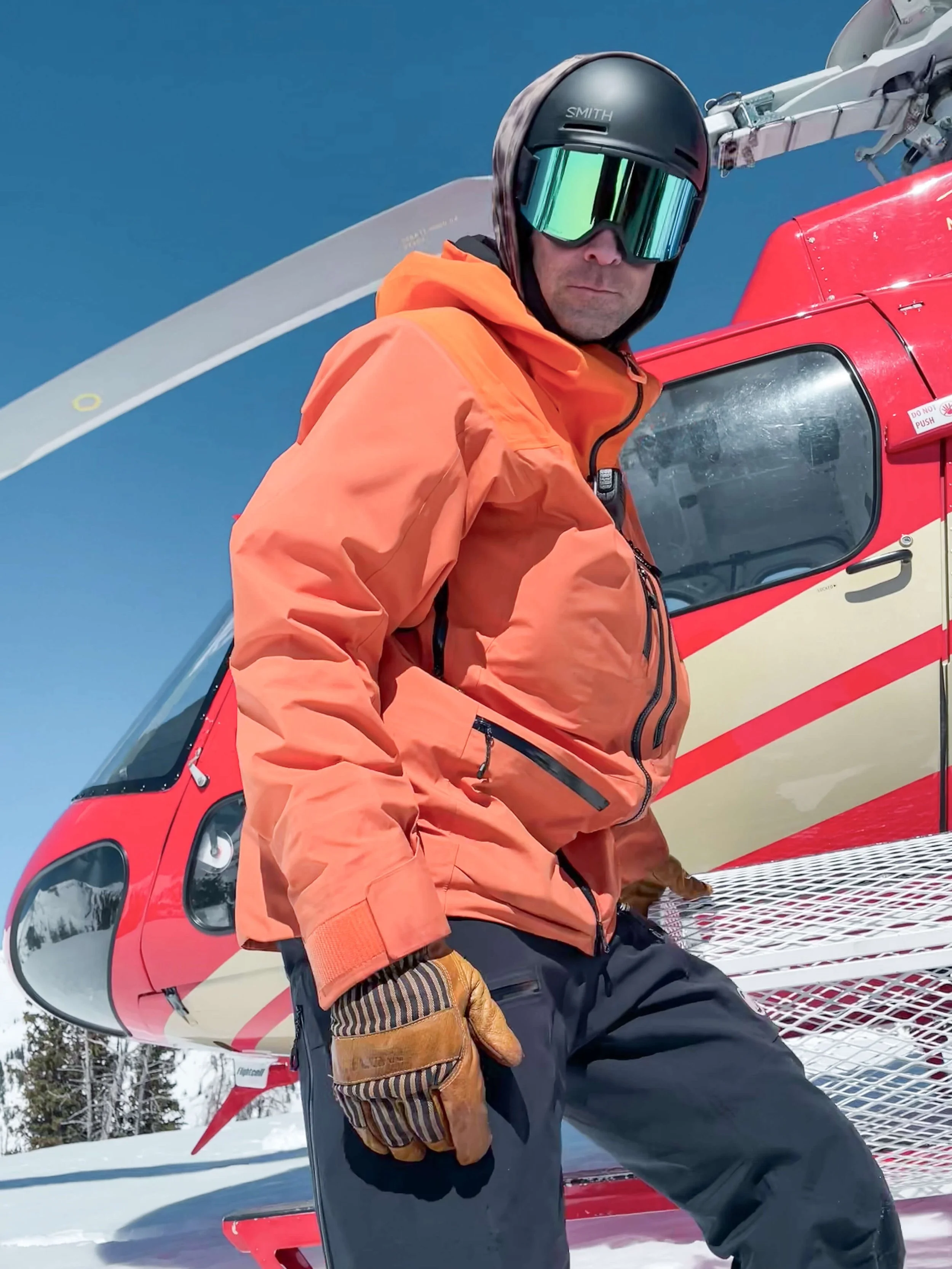 A person dressed in orange winter gear, wearing a black helmet and reflective ski goggles, sitting on a red helicopter outdoors in a snowy environment.