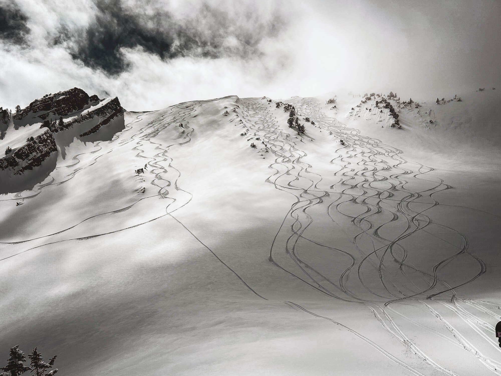 Snow-covered mountain slope with numerous ski or snowboard tracks. Some trees are visible on the slope, and clouds cover part of the sky.