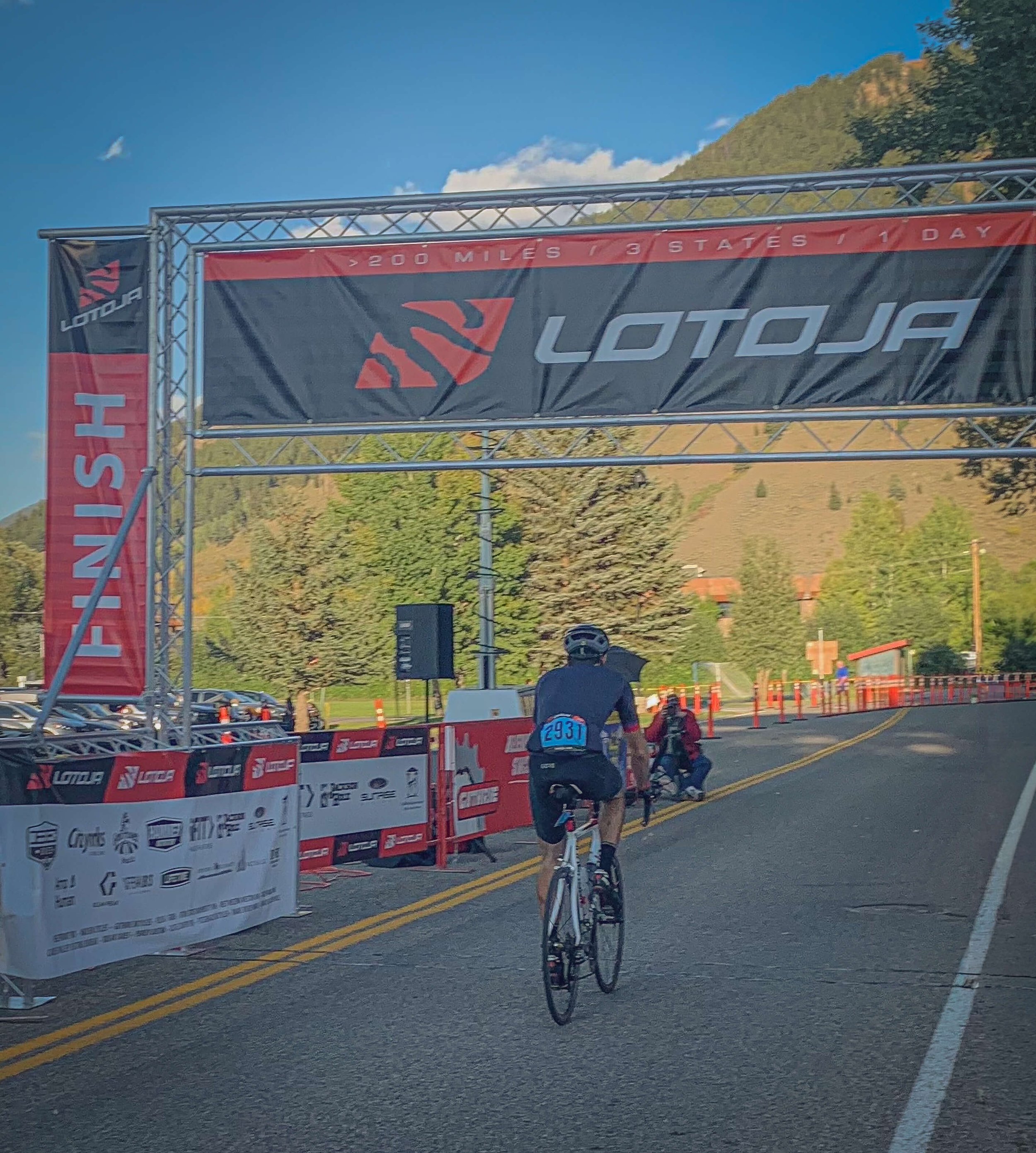Cyclists crossing the finish line at the LOTOJA race, with a large banner overhead that reads ' 200 MILES | 3 STATES | 1 DAY' and a backdrop of mountains and trees.