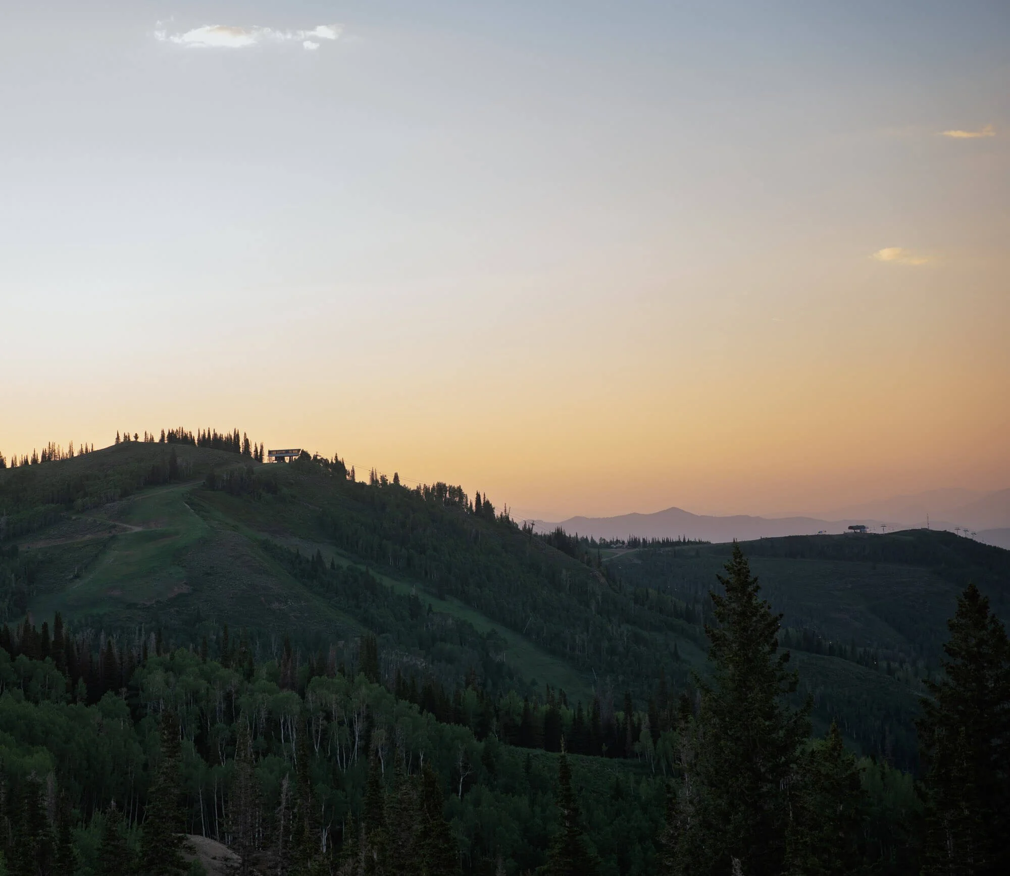 Landscape of green rolling hills with trees and a small building on top, during sunset with a sky transitioning from light blue to orange.