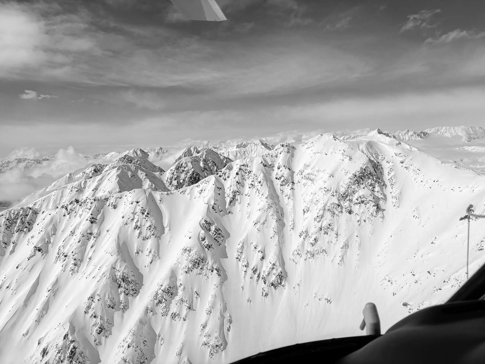 Black and white photo of snow-covered mountain peaks taken from inside a plane, with part of the plane's windshield and dashboard visible in the foreground.