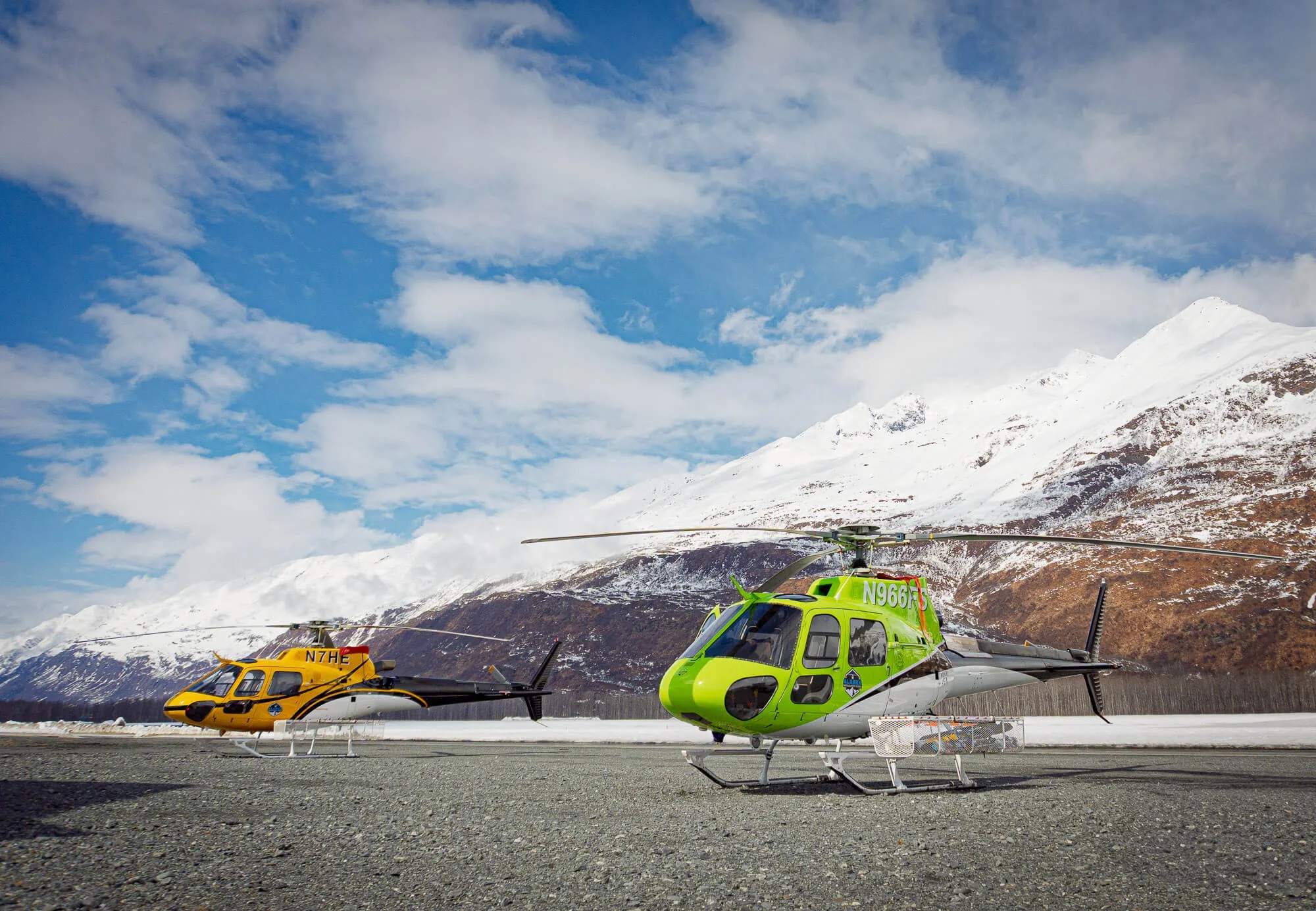 Two helicopters, one green and one yellow, parked on a flat surface with snow-covered mountains in the background under a partly cloudy sky.