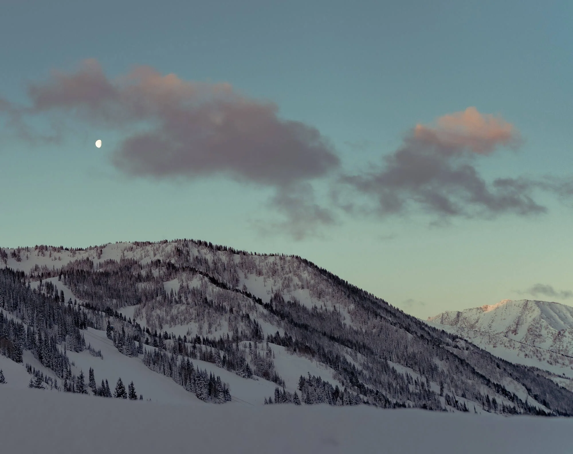 Snow-covered mountain range under a sky with clouds and a visible moon.