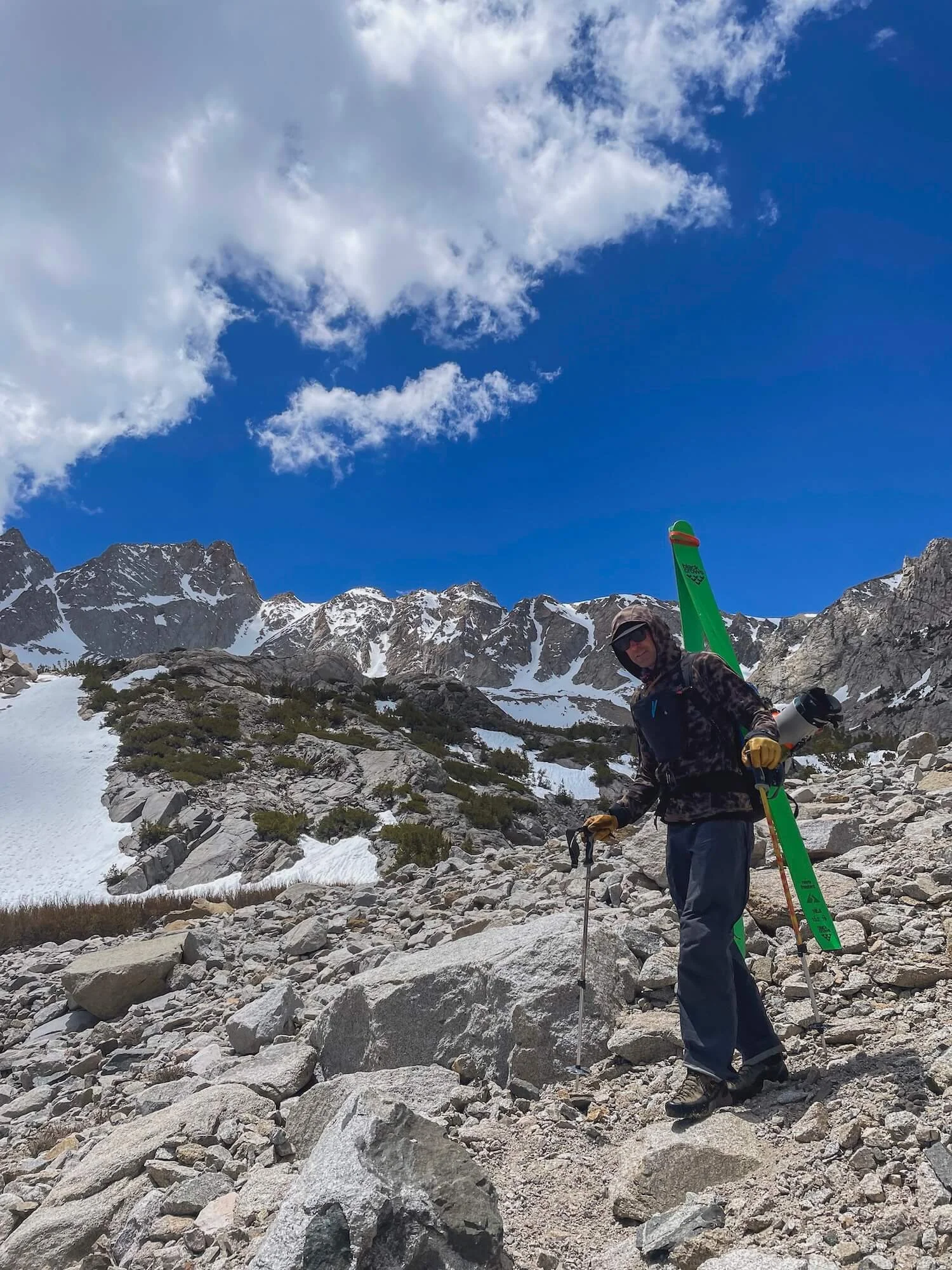 A person hiking on rocky terrain in a mountainous area with snow patches, carrying a green sled or snowboard on their back, wearing sunglasses, a hooded jacket, and gloves, under a blue sky with scattered clouds.