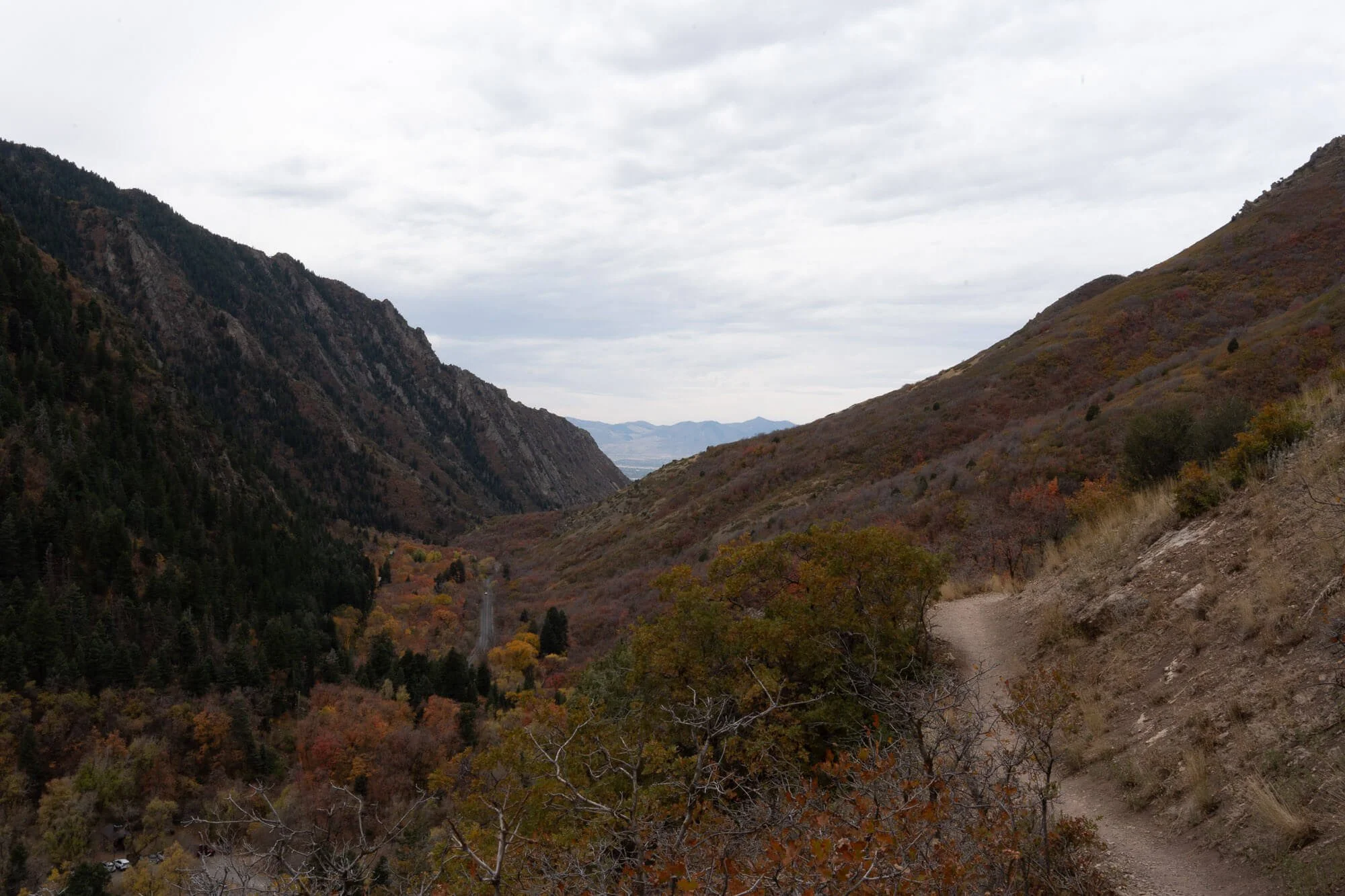 A mountain valley with a dirt trail on the right side, surrounded by autumn-colored trees and hills under a cloudy sky.