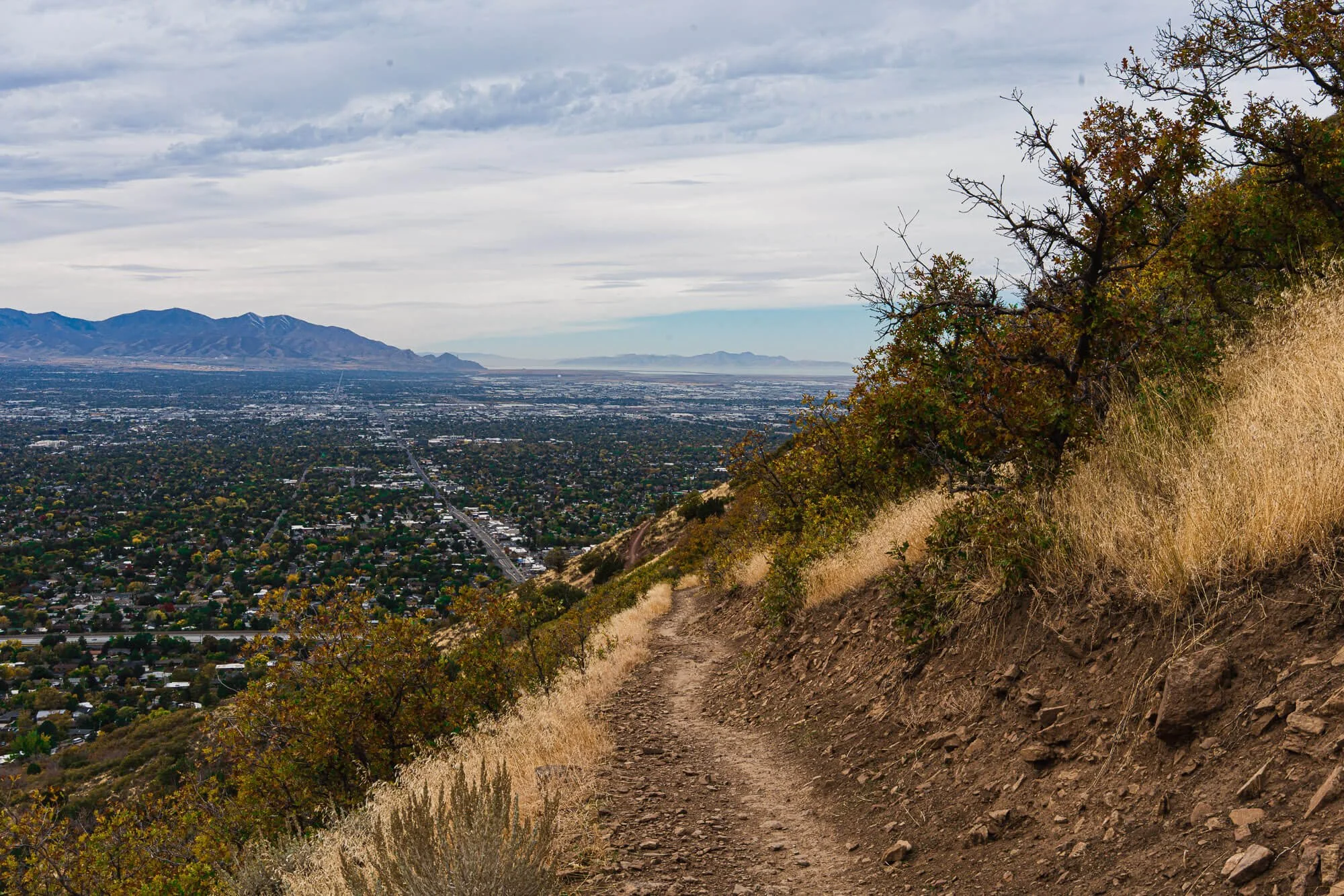 A dirt hiking trail on a hillside with dry grass and small shrubs, overlooking a city with mountains in the background under cloudy skies.