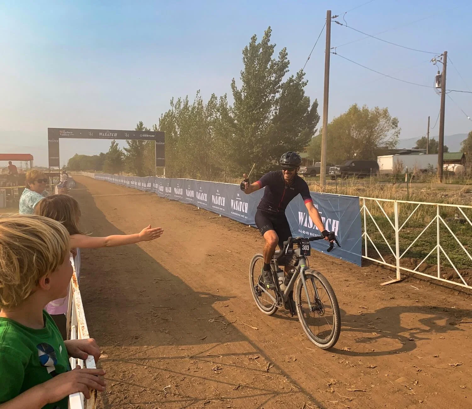 A cyclist in black gear riding a mountain bike on a dirt trail, giving a thumbs-up, as spectators cheer from the side of the trail near a finish line in an outdoor race setting.