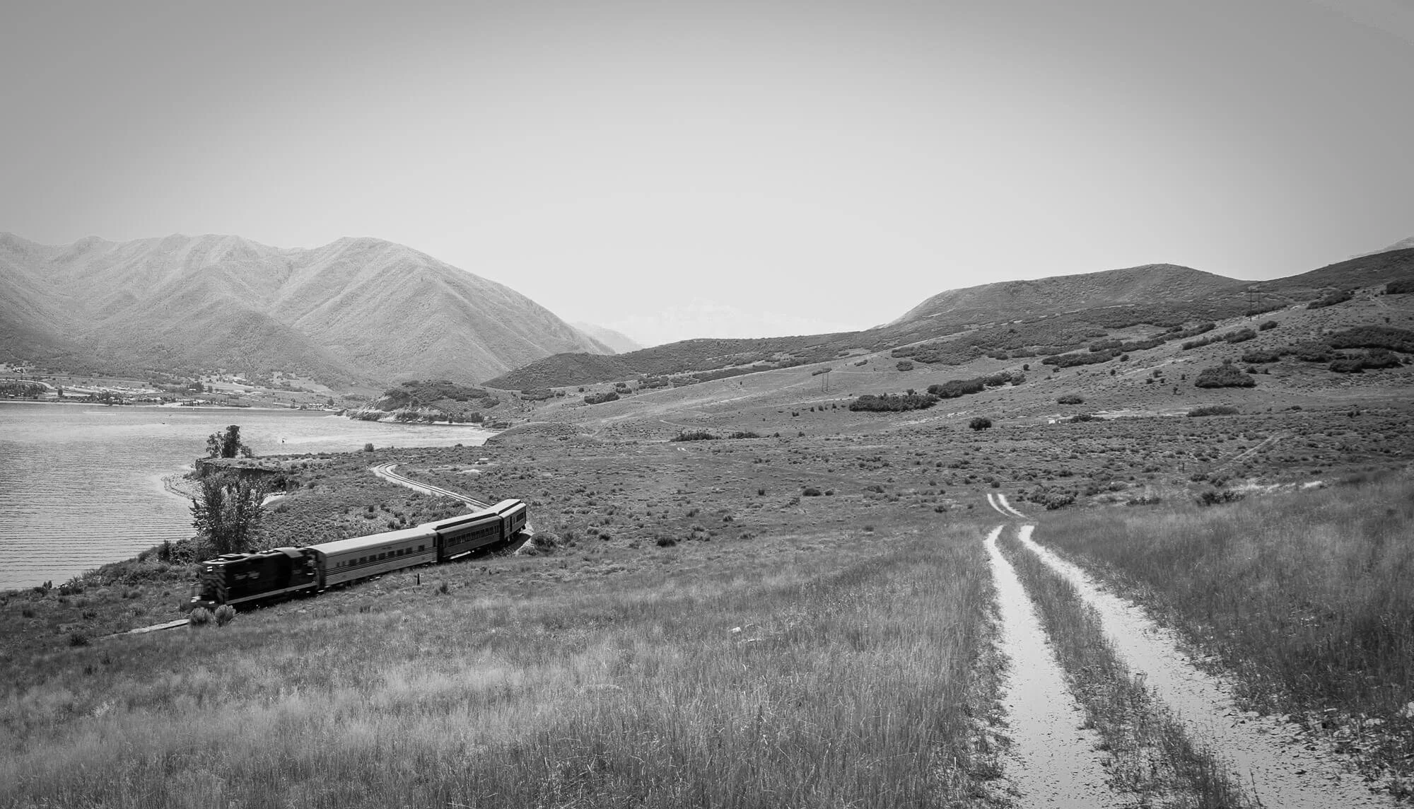 A black and white landscape with a train traveling along a curved track near a lake, surrounded by hills and mountains.