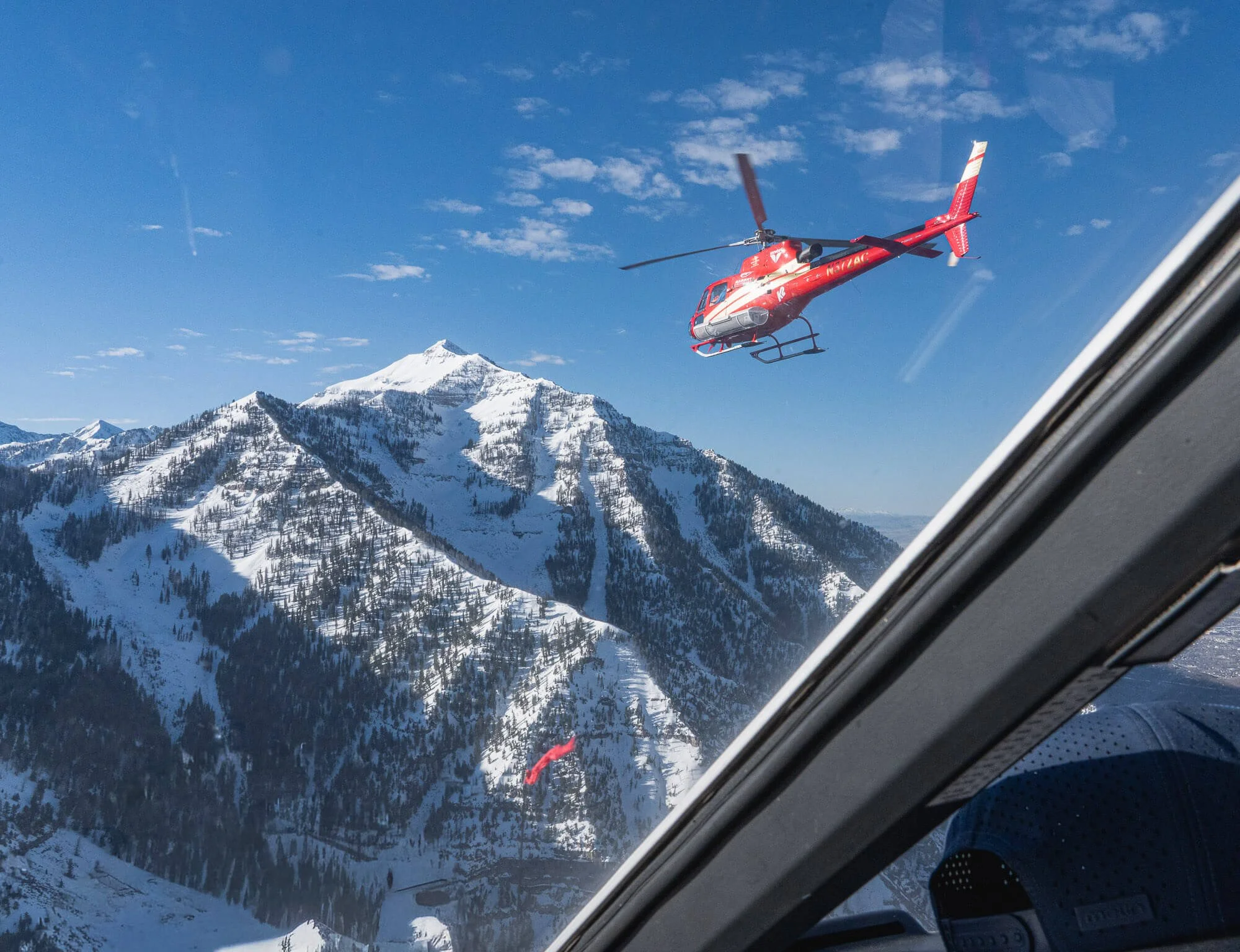 A helicopter flying over snowy mountain peaks on a clear day, seen from inside an aircraft.