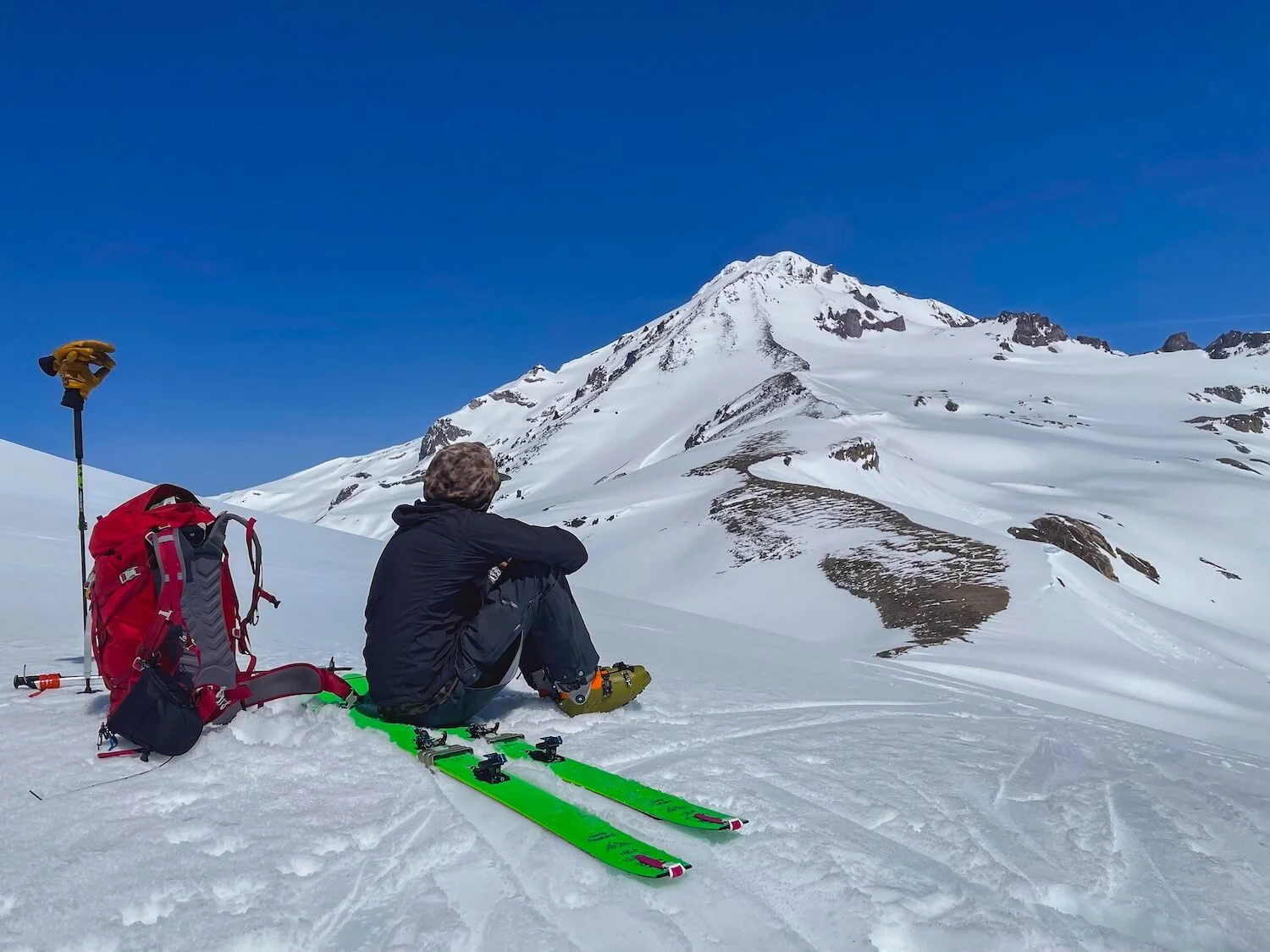 A person sitting on snow with green skis, a red backpack, and ski poles nearby, overlooking snow-covered mountains and a clear blue sky.