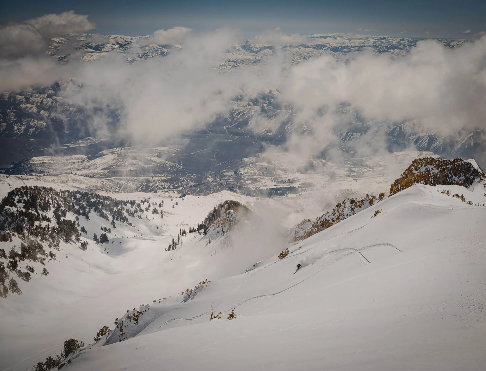 Snow-covered mountain slope with trees and distant valley, partly obscured by clouds.