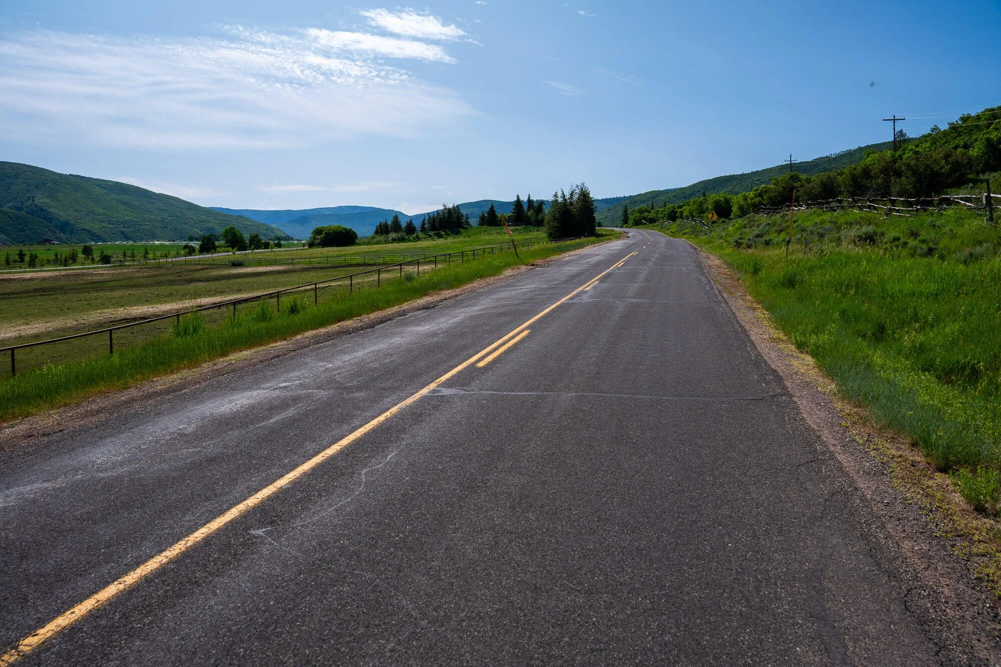 A rural asphalt road stretching through green fields and rolling hills, under a partly cloudy blue sky.