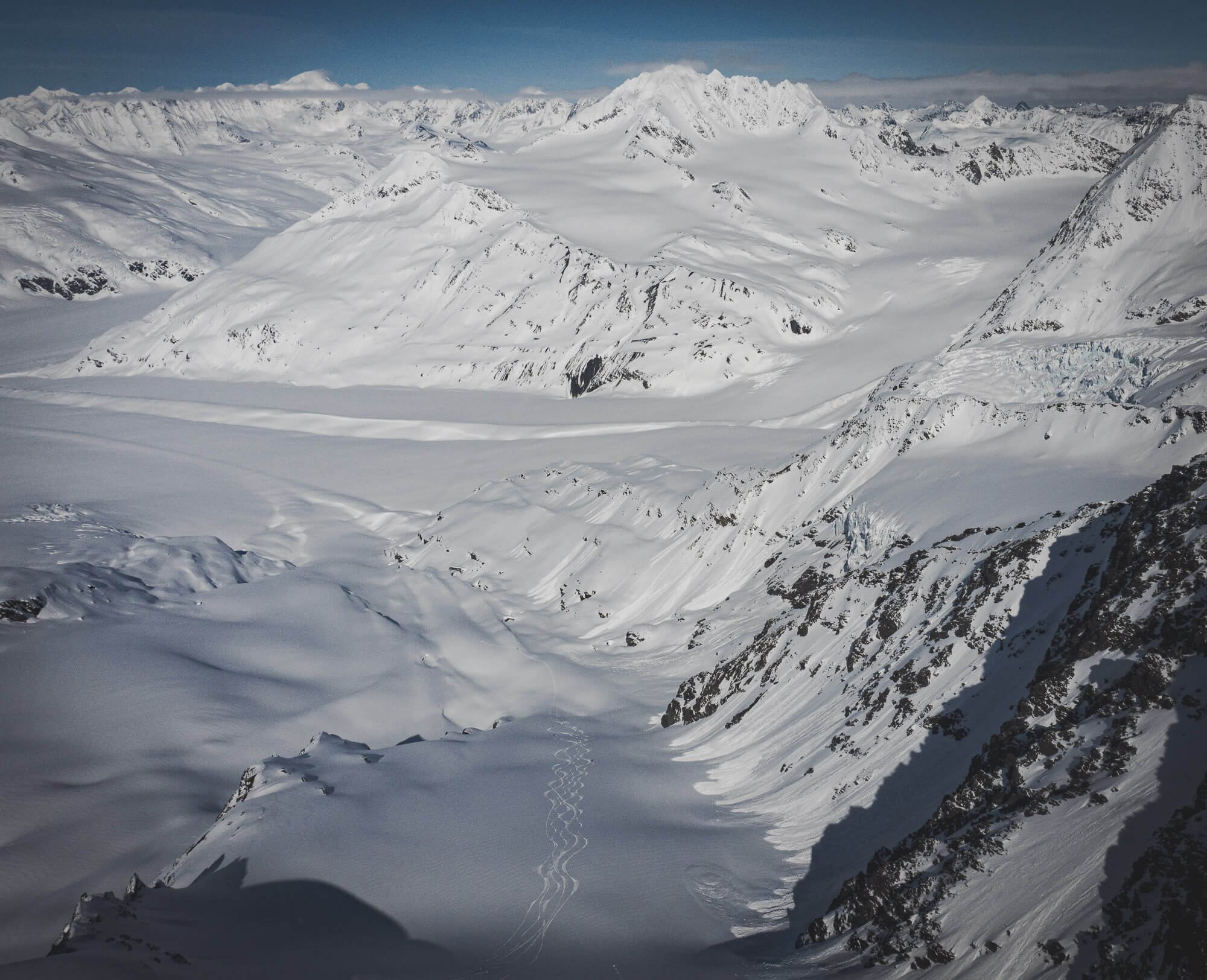 Snow-covered mountain landscape with glacier, steep rocky cliffs, and mountain peaks in the distance under a clear sky.