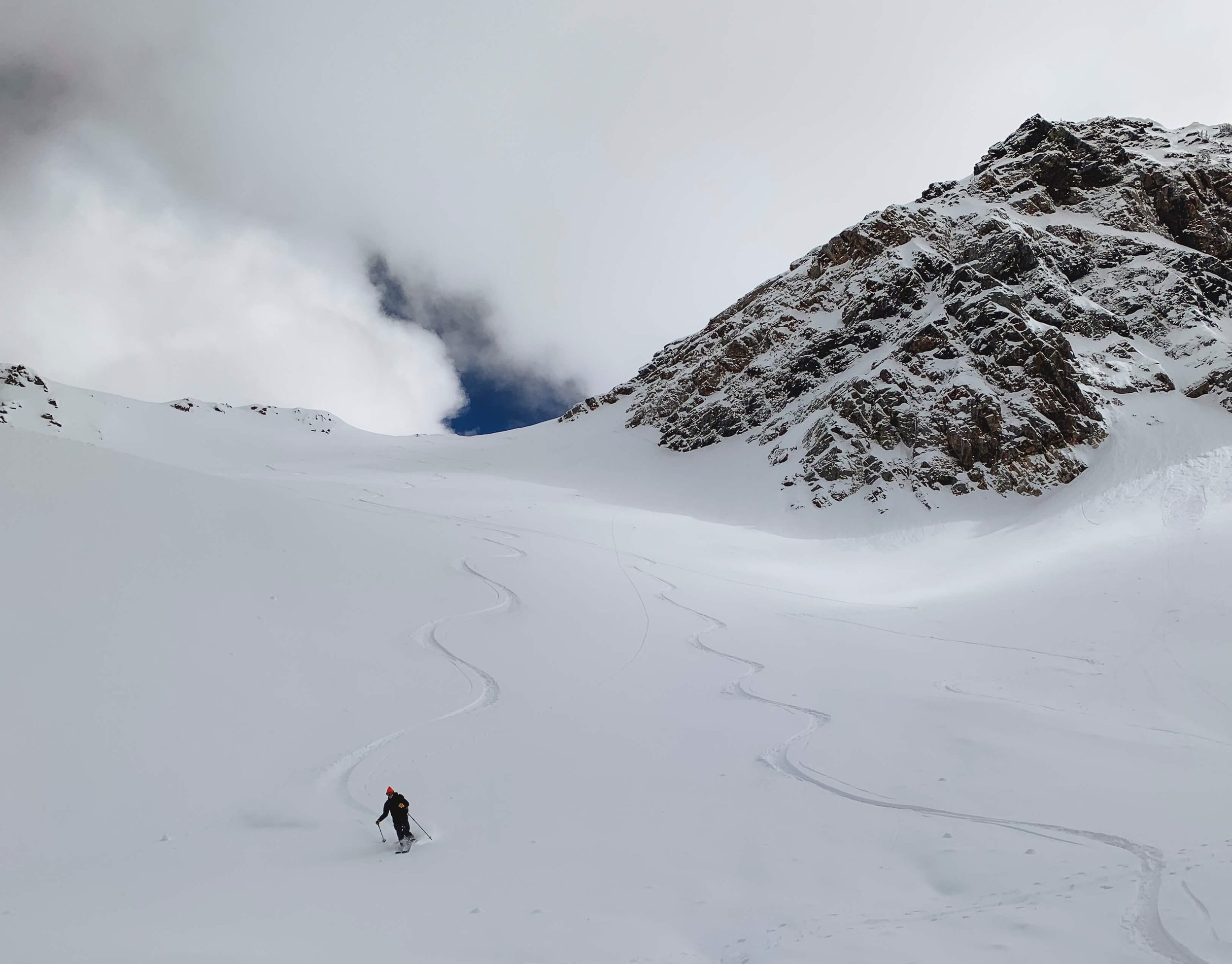 A skier climbing a snow-covered mountain slope with a mountain and cloudy sky in the background.