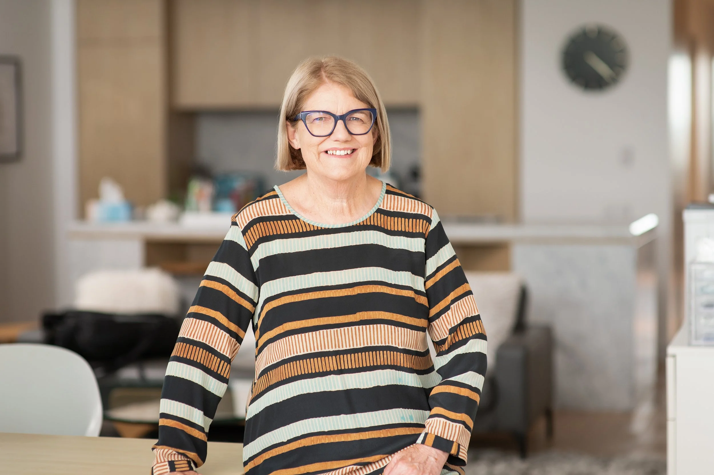 A smiling woman with glasses standing indoors in a home setting.