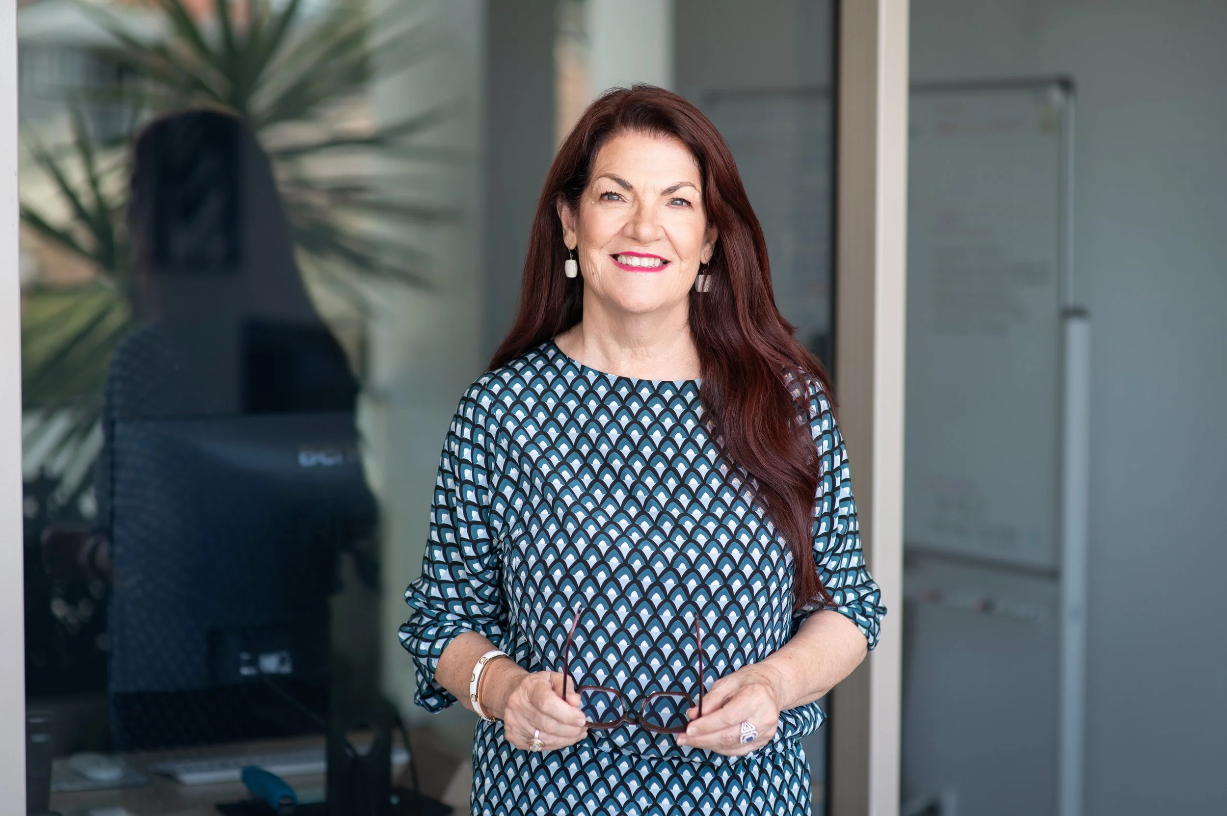 A woman with long red hair, wearing a blue patterned dress, standing indoors near a glass door holding her glasses. She is smiling.