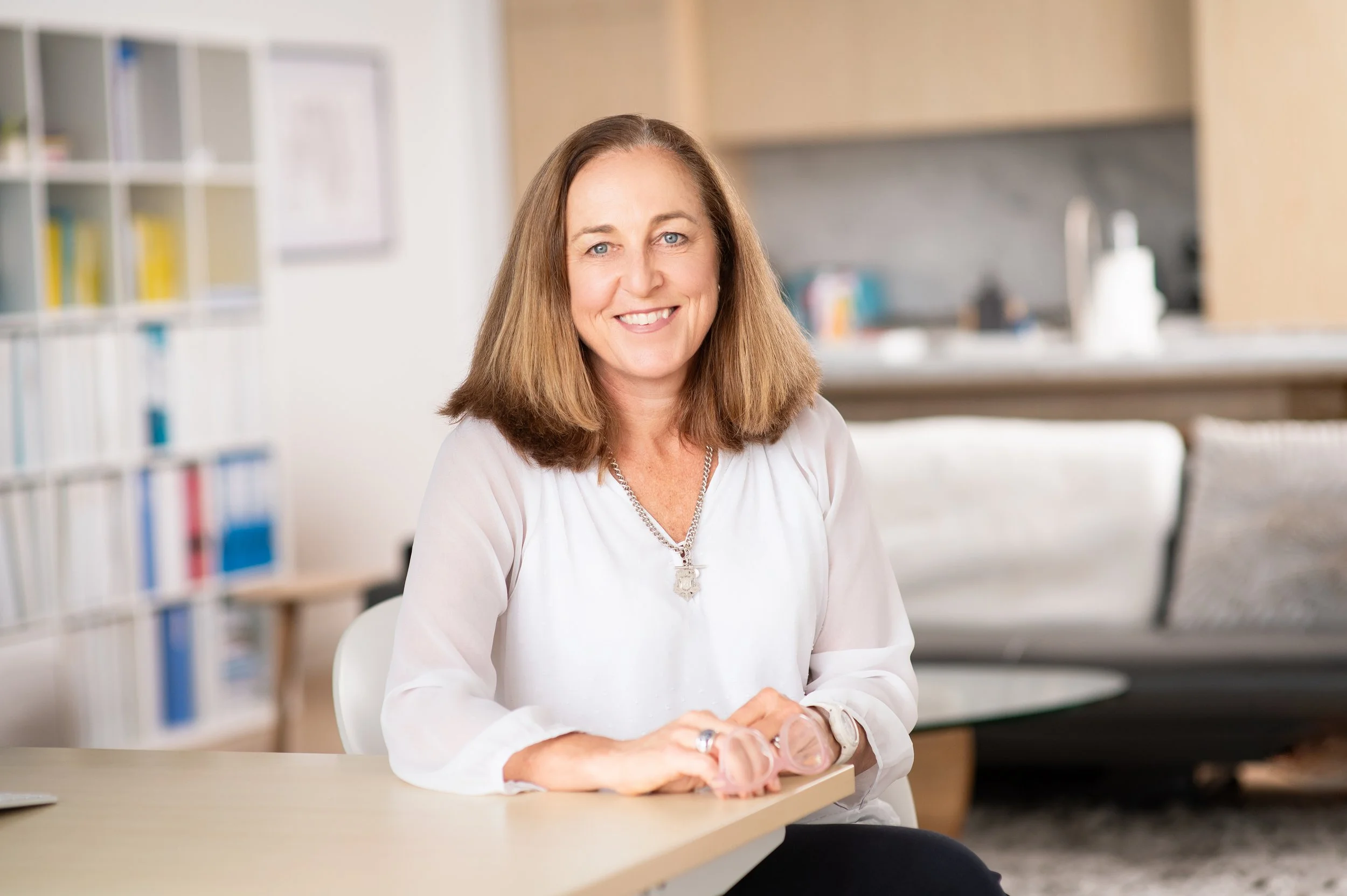 A woman with shoulder-length brown hair and a white blouse smiling, sitting at a table in a modern living room or office space.