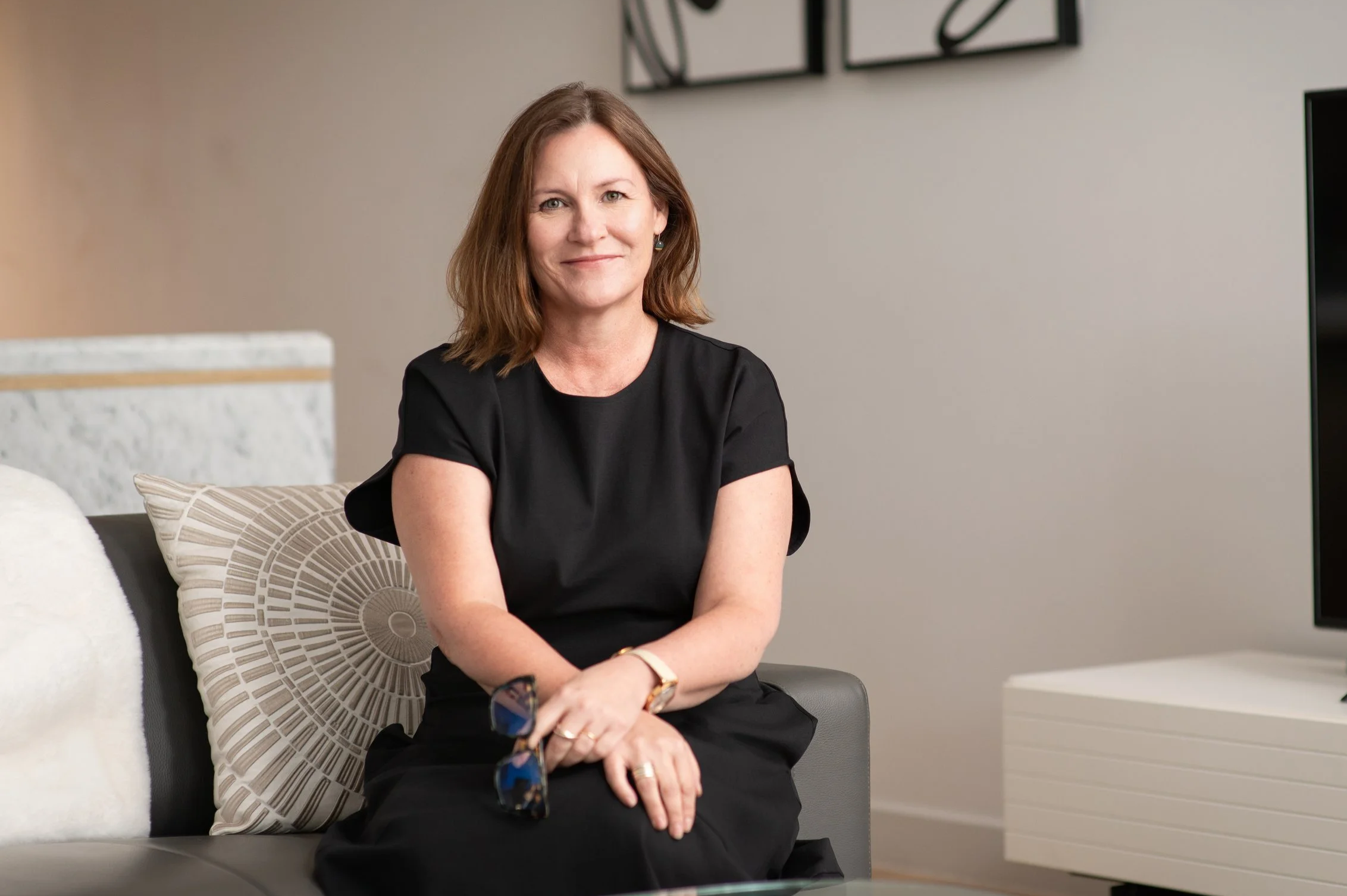 A woman with shoulder-length brown hair, wearing a black dress, sitting on a gray sofa in a modern room, smiling at the camera.