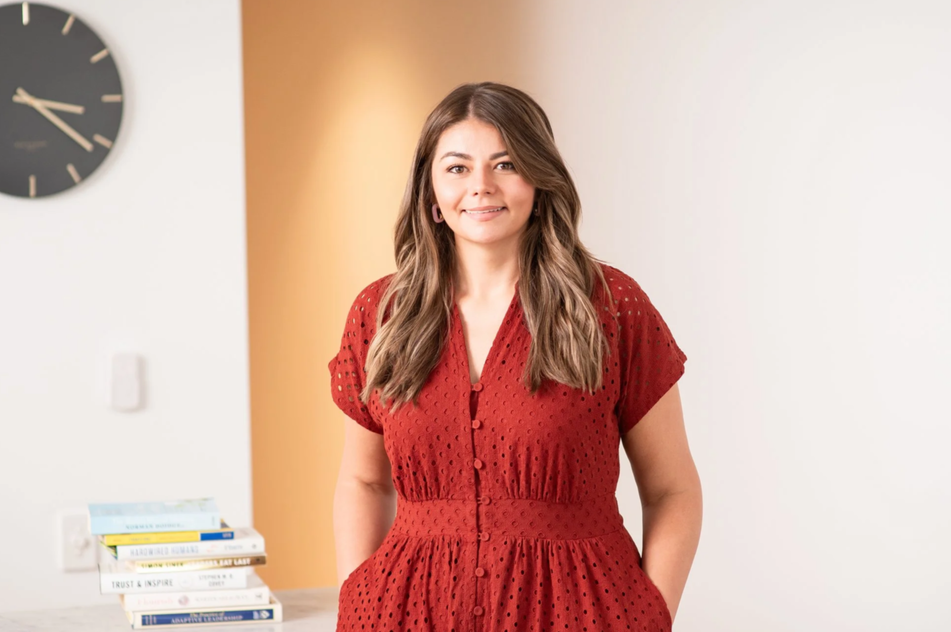 Young woman in red dress smiling indoors with books and a wall clock in background.