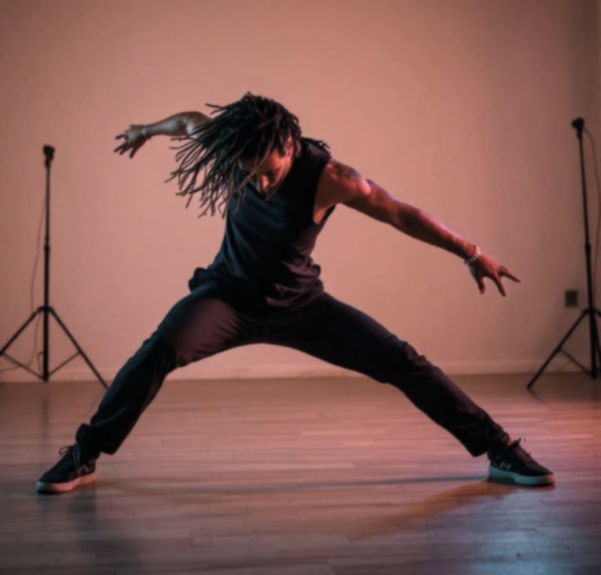 A person dancing in a studio with a pink and orange background, wearing a black tank top, black pants, and sneakers, with dreadlocks and tattoos, in a wide-legged pose.