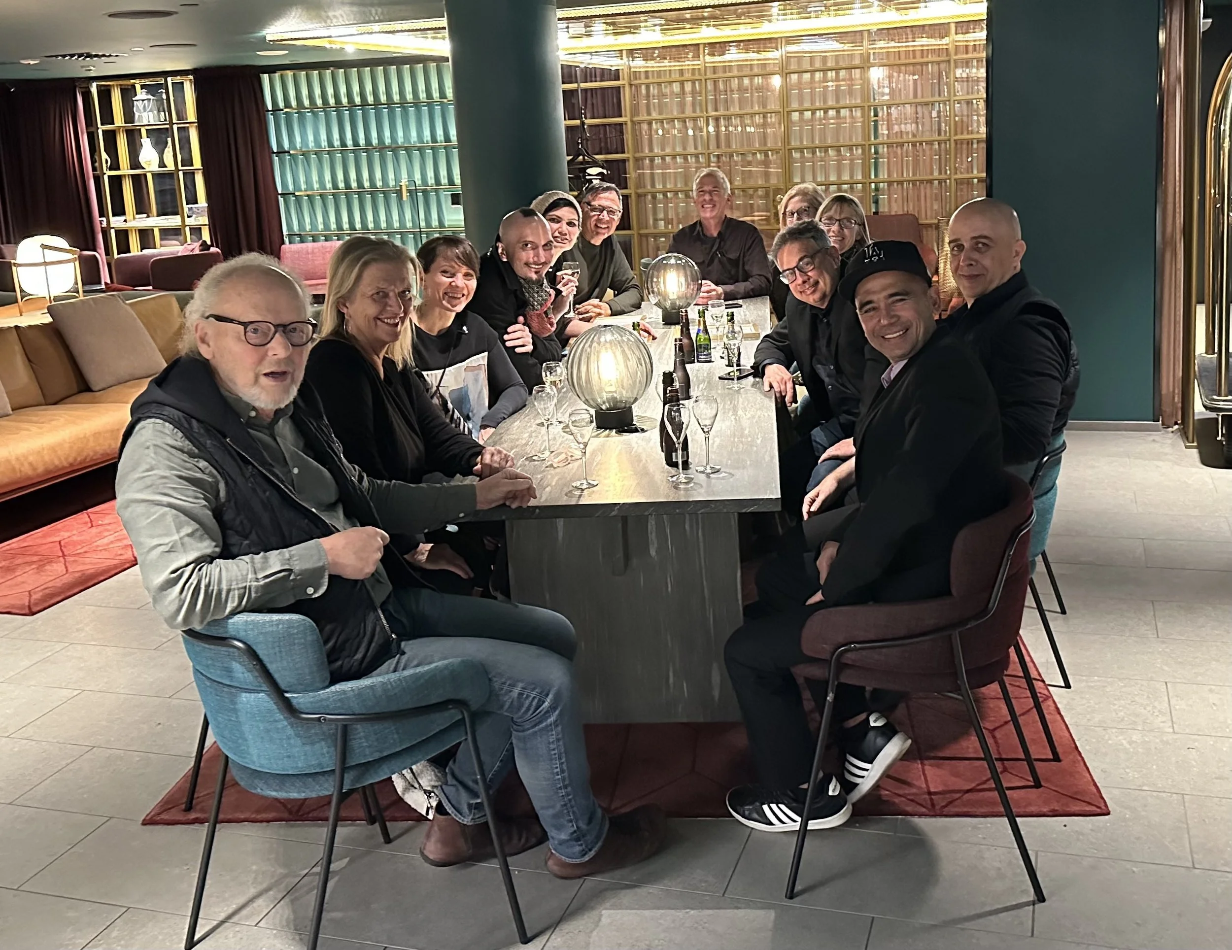 A group of fourteen people sitting around a long dining table in a stylish restaurant, smiling and posing for the photo, with drinks and glasses on the table.