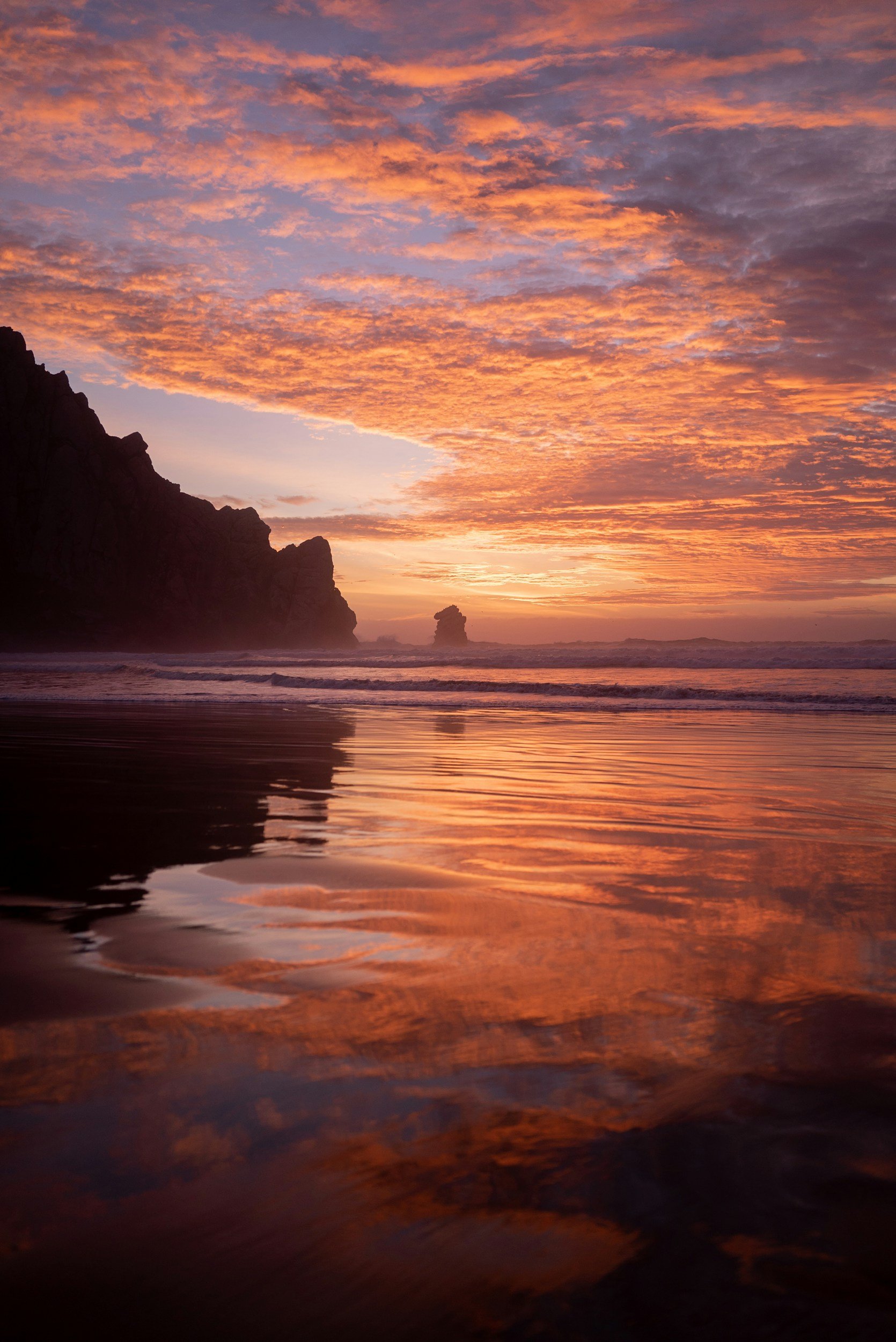 Sunset over the ocean with colored clouds, dark cliffs on the left, and the reflection of the sky on wet sand in the foreground.