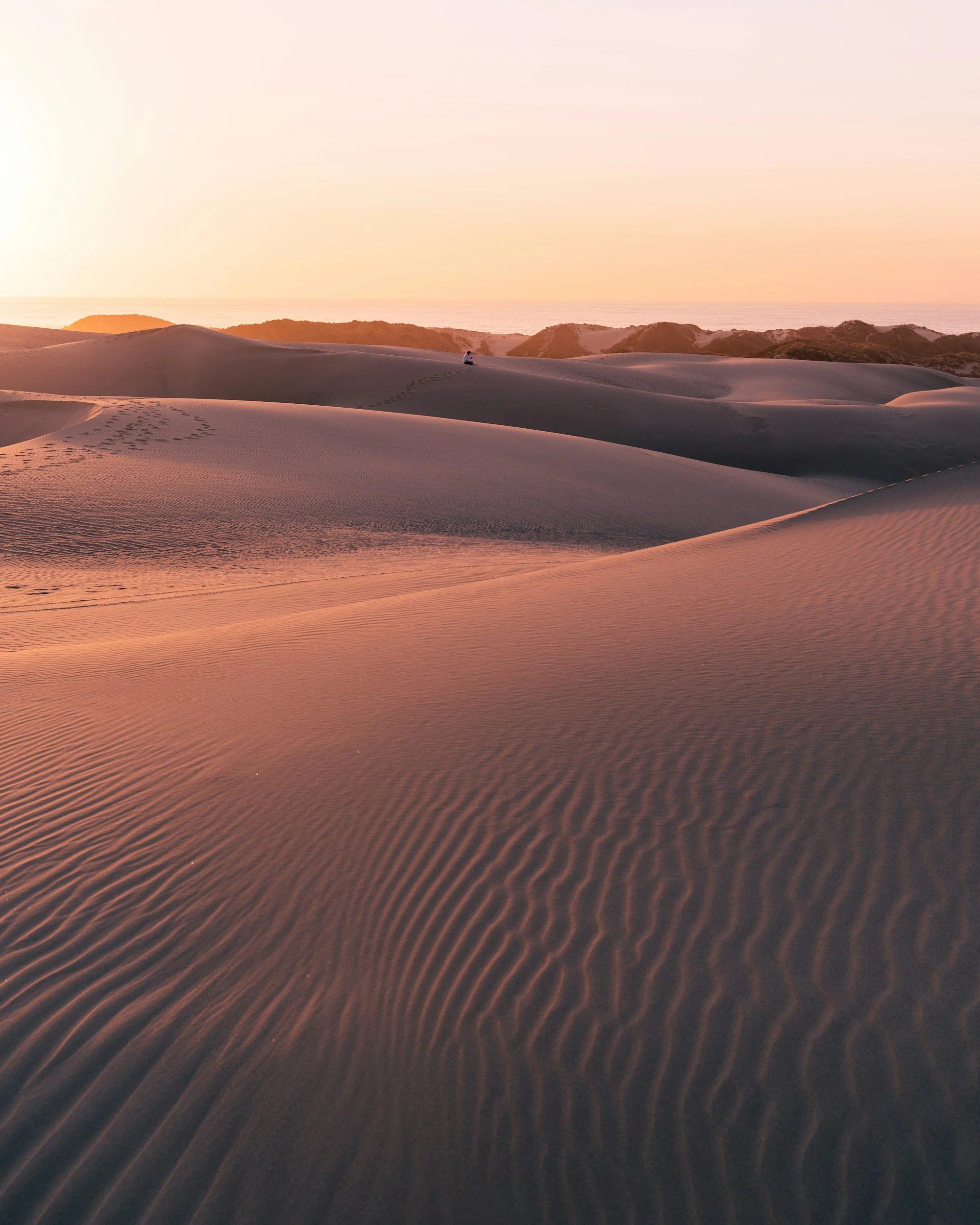 Sand dunes at sunset with footprints and a person sitting in the distance.