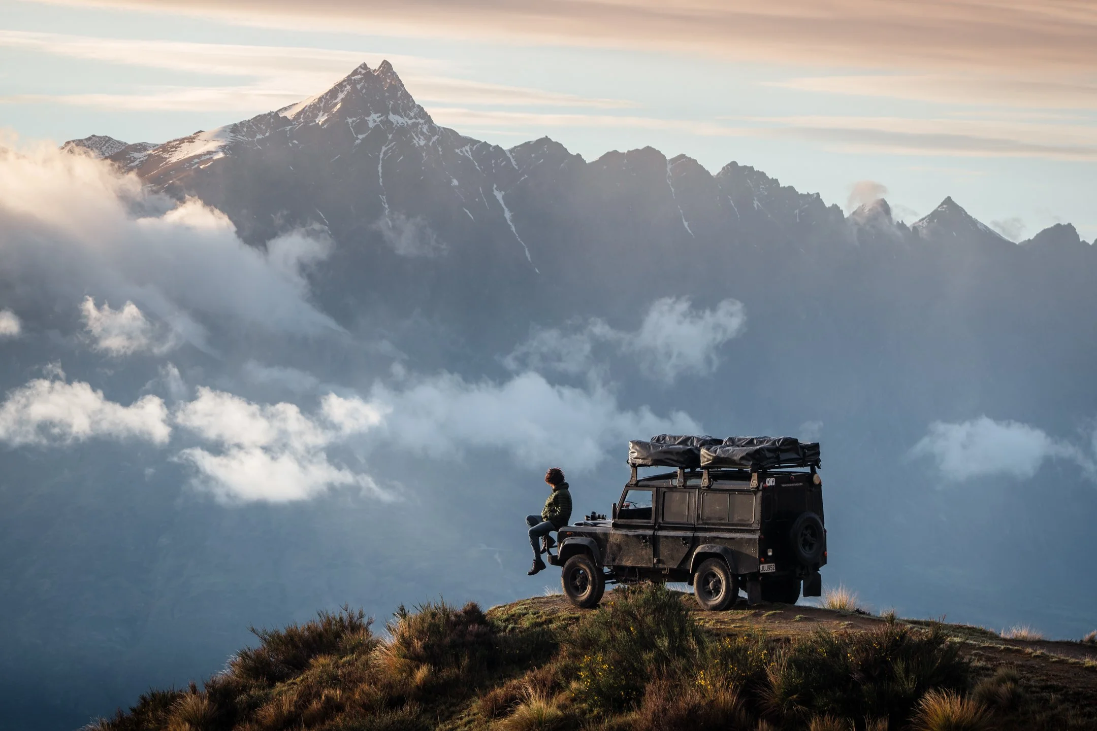 Two people sitting on the back of a black off-road vehicle parked on a grassy hilltop, overlooking a mountain range with snow-capped peaks and clouds at sunrise or sunset.