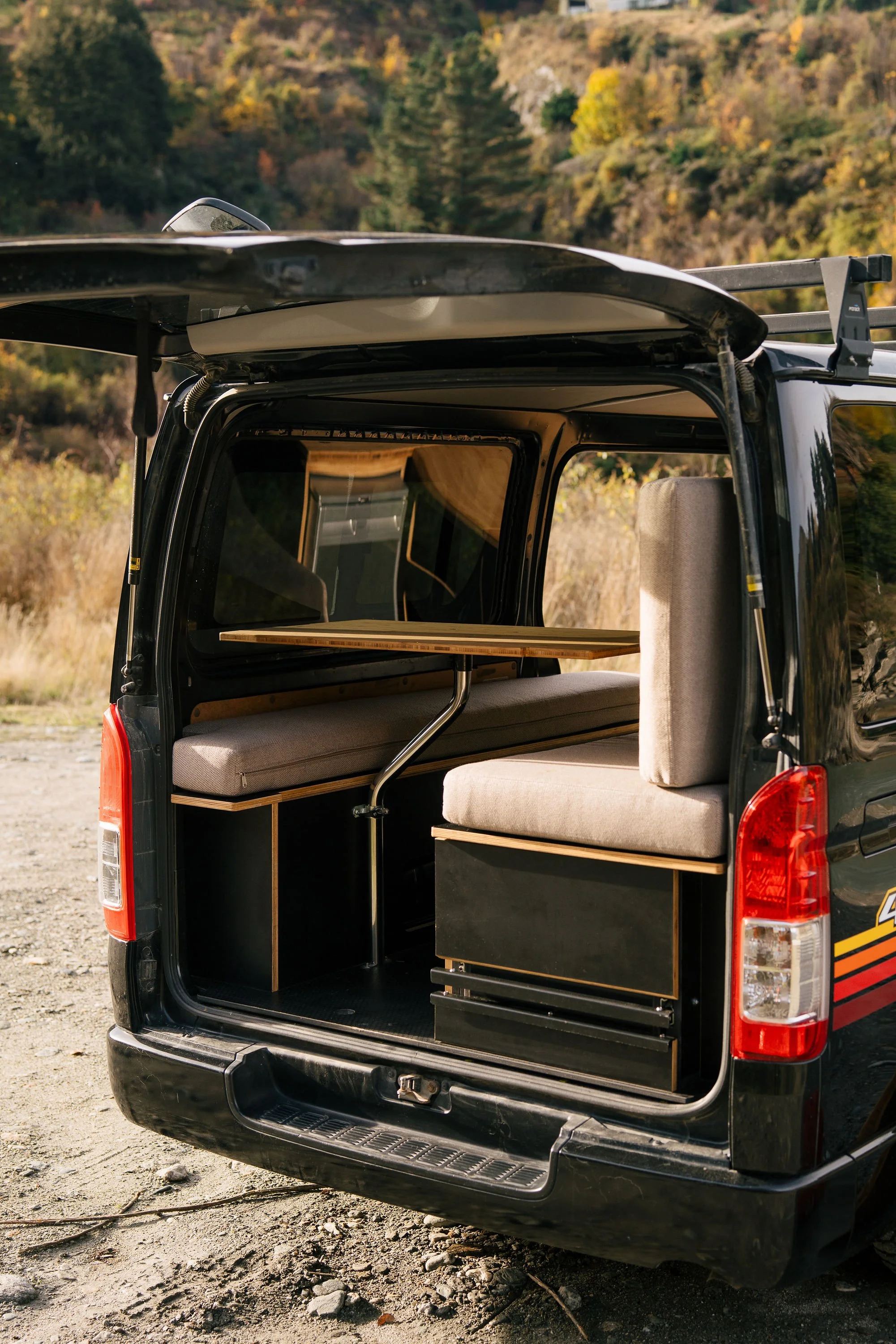 The back of a black van with its rear door open, revealing a built-in seating and storage area with a wooden table inside, set against a backdrop of hills covered with autumn trees.