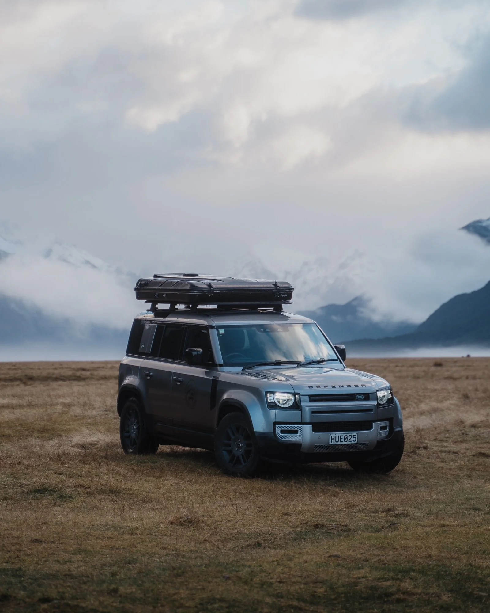A silver SUV with a roof box parked on a grassy field, mountains in the background and cloudy sky above.
