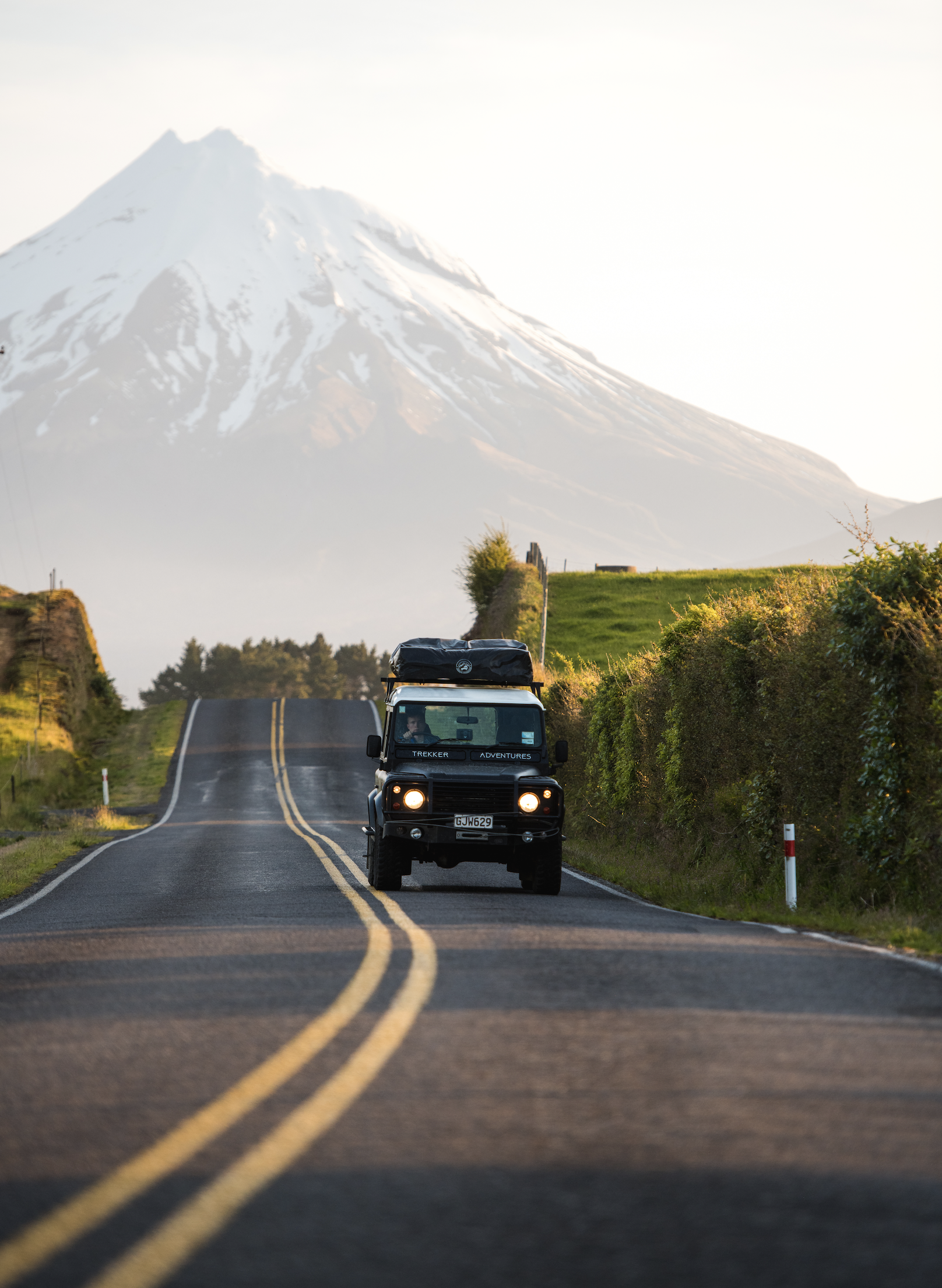 A black off-road vehicle driving down a two-lane road with a double yellow line, surrounded by greenery, with a snow-capped mountain in the background.