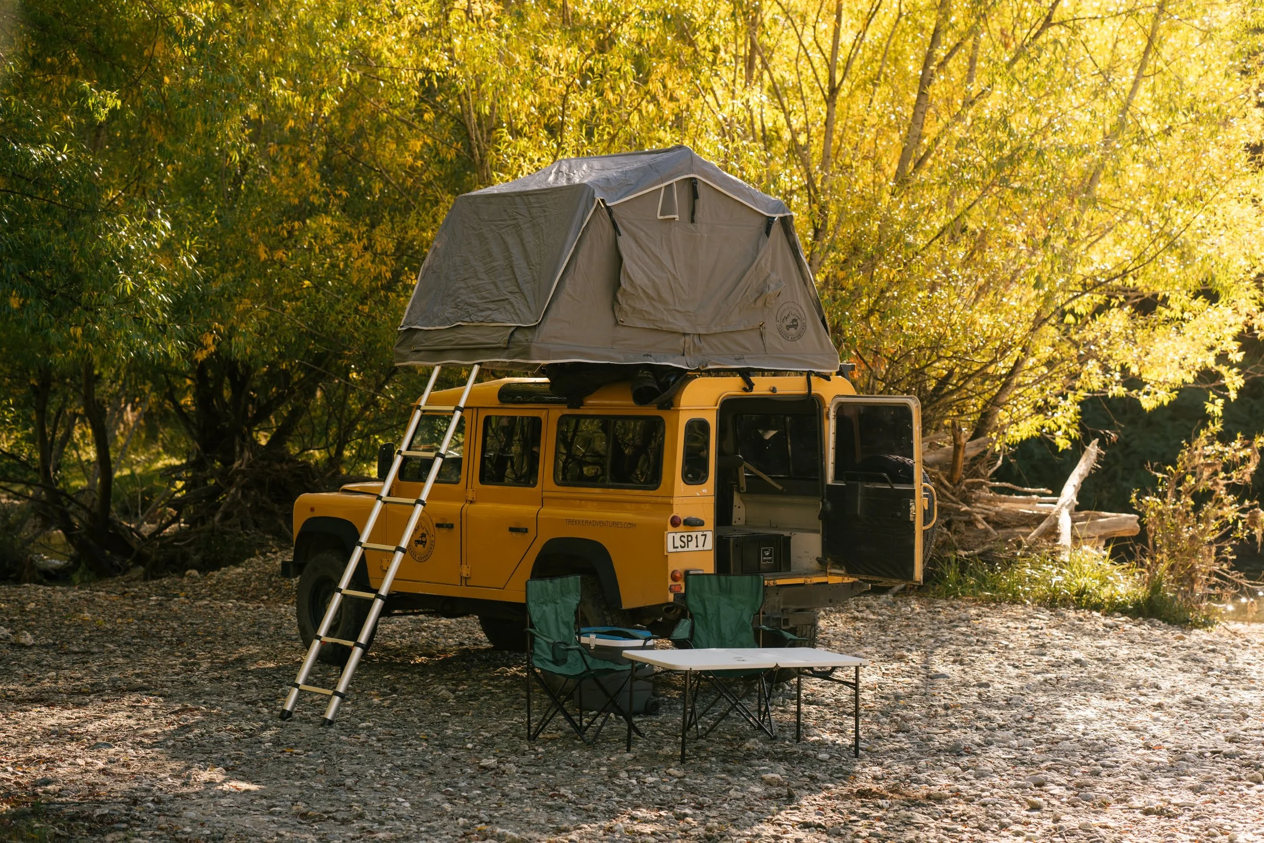 Yellow off-road vehicle with a rooftop tent, a ladder, and camping chairs set up in a wooded area with autumn leaves.