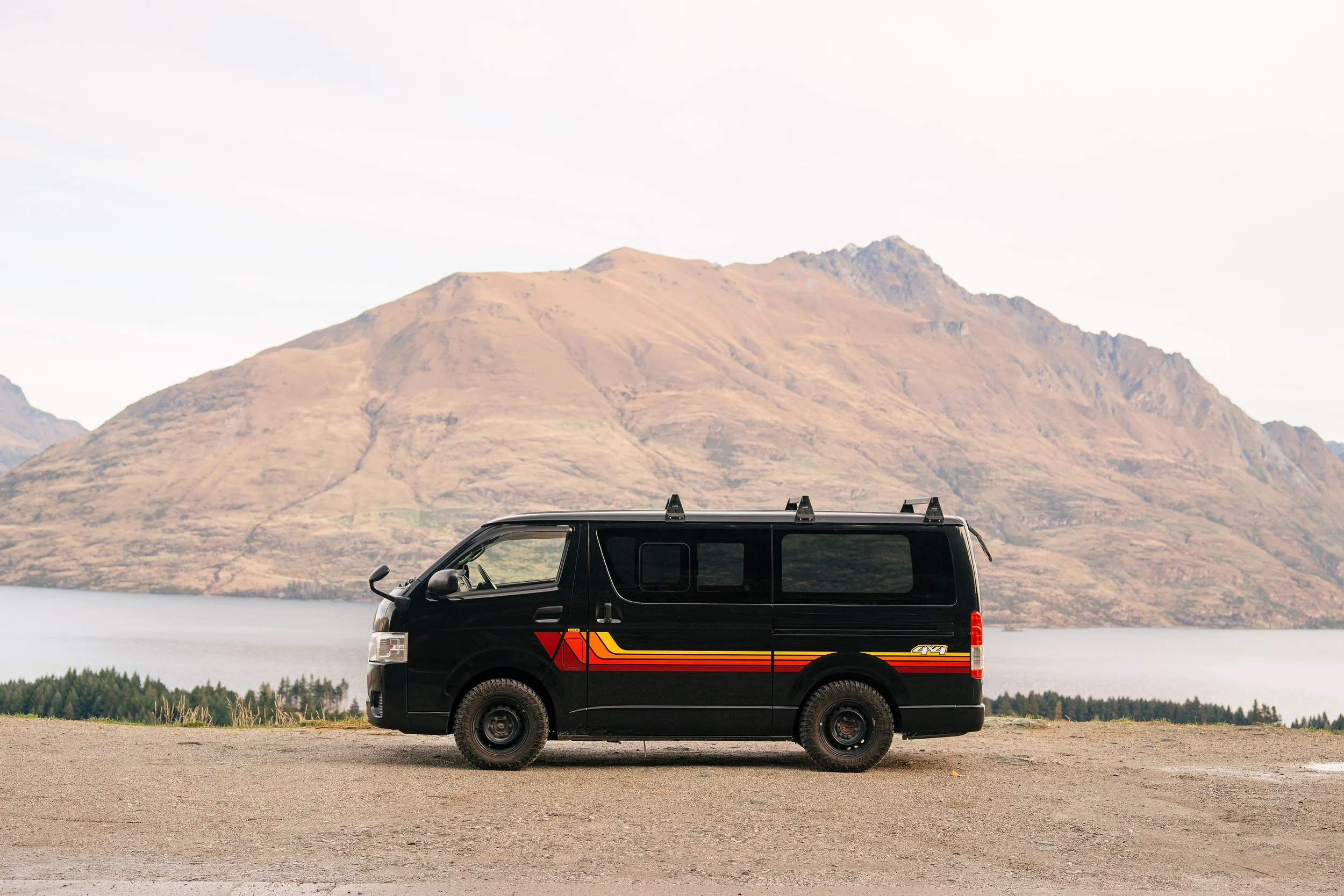 A black van with colorful stripes parked near a body of water, with mountainous terrain in the background under a cloudy sky.