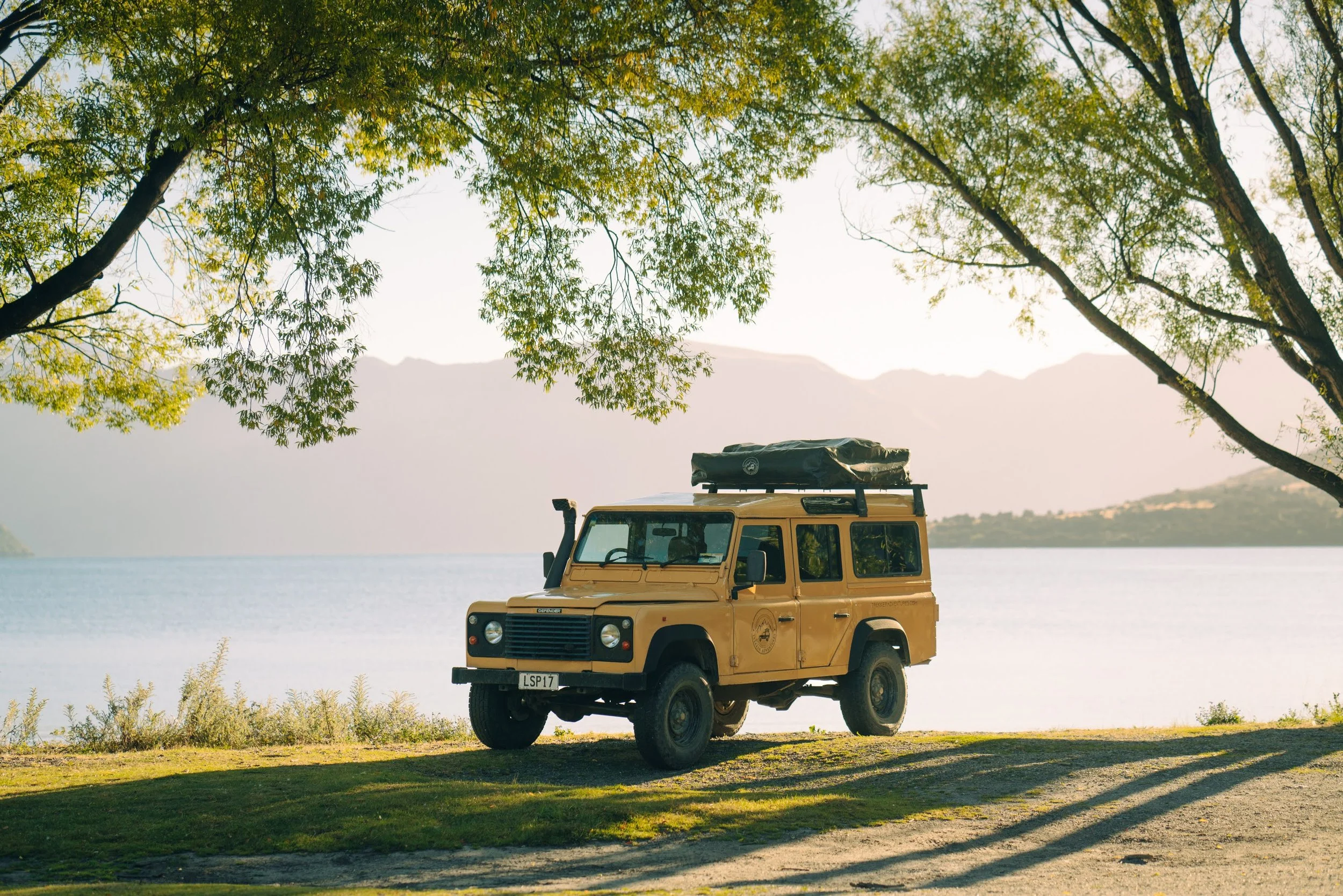 Yellow Land Rover Defender parked near a lake, with green trees overhead and mountains in the background during daytime.