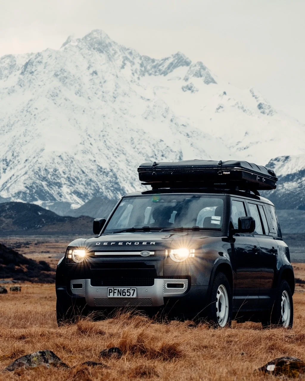 A black Land Rover Defender parked on a grassy field with a mountain range in the background, snow-covered peaks and a cloudy sky.