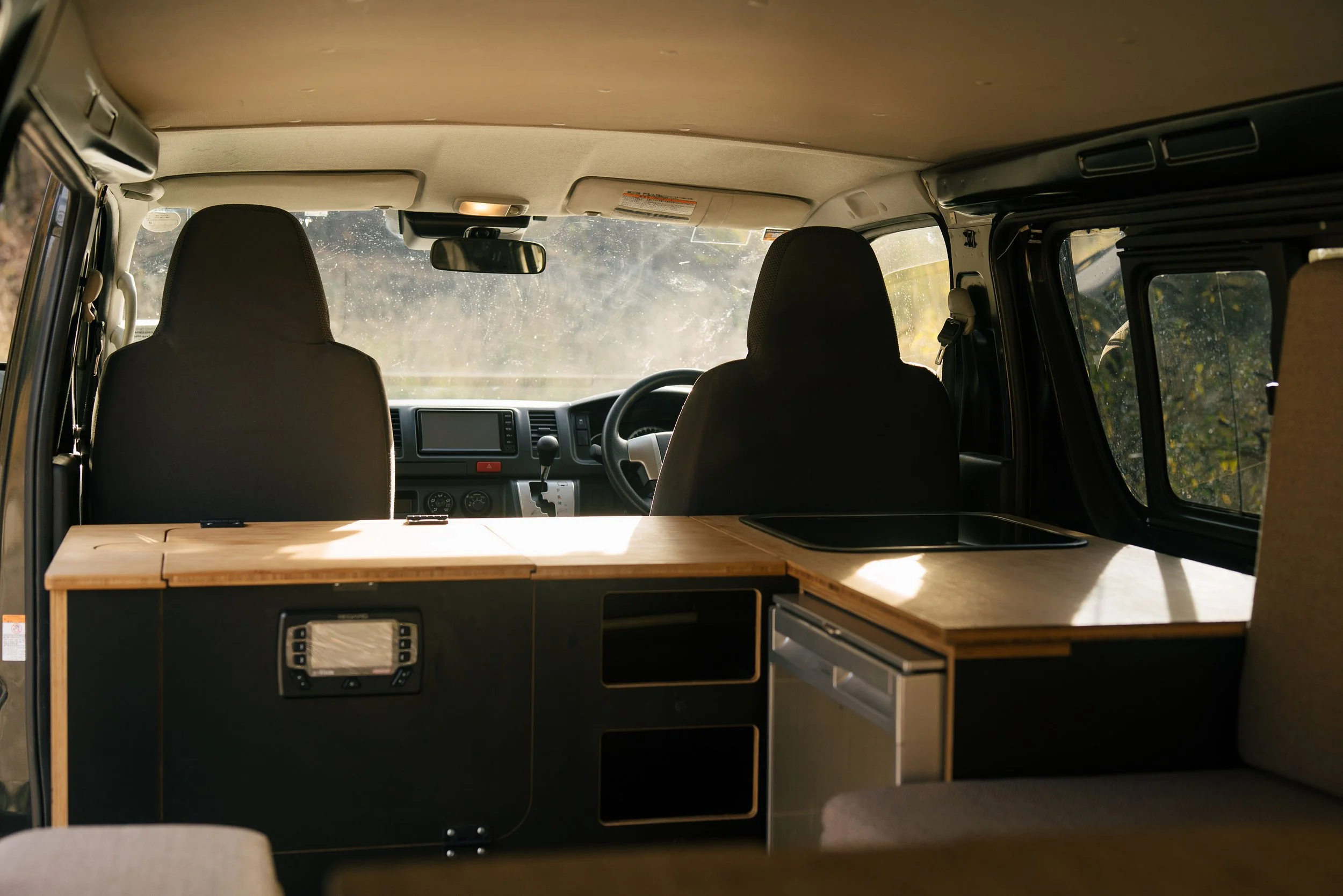 Inside view of a van with front seats, a dashboard, and a kitchenette area with wooden countertops and drawers, sunlight coming through the windows.