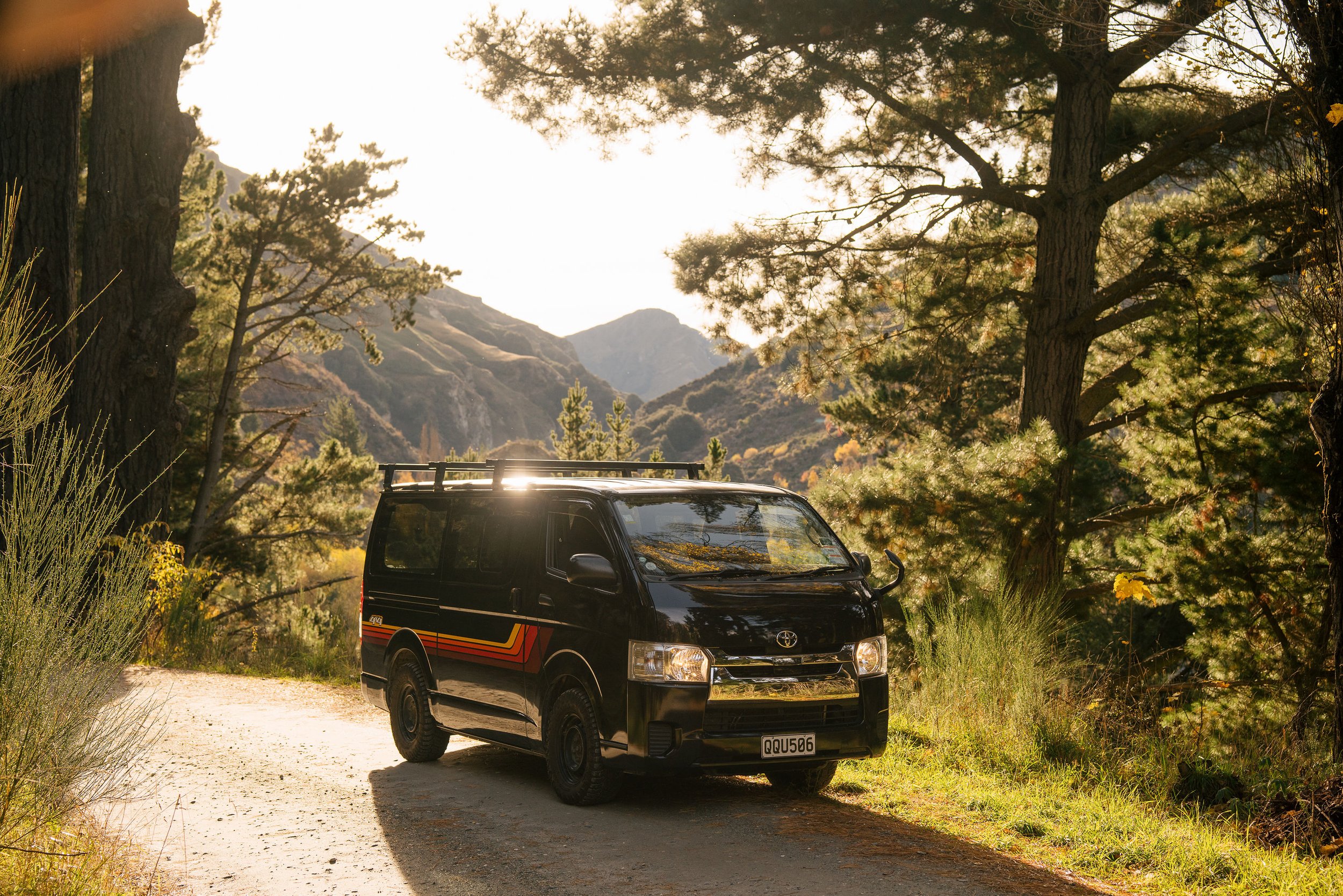 A black van with a roof rack parked on a dirt road surrounded by tall trees and mountains in the background, with sunlight filtering through the branches.