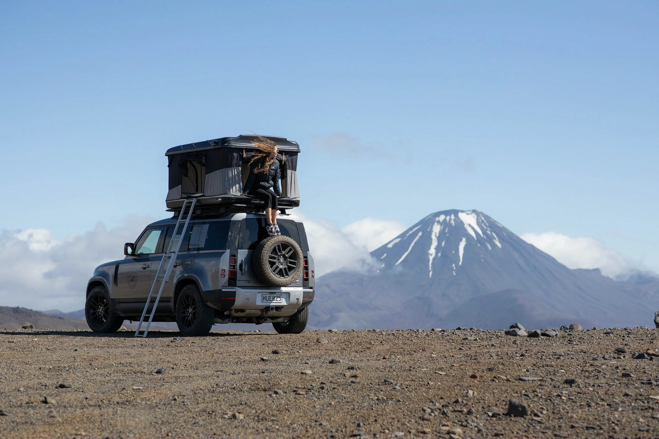 A person sitting on top of a black off-road vehicle with a rooftop tent, in a barren landscape with Mount Fuji in the background under a partly cloudy sky.
