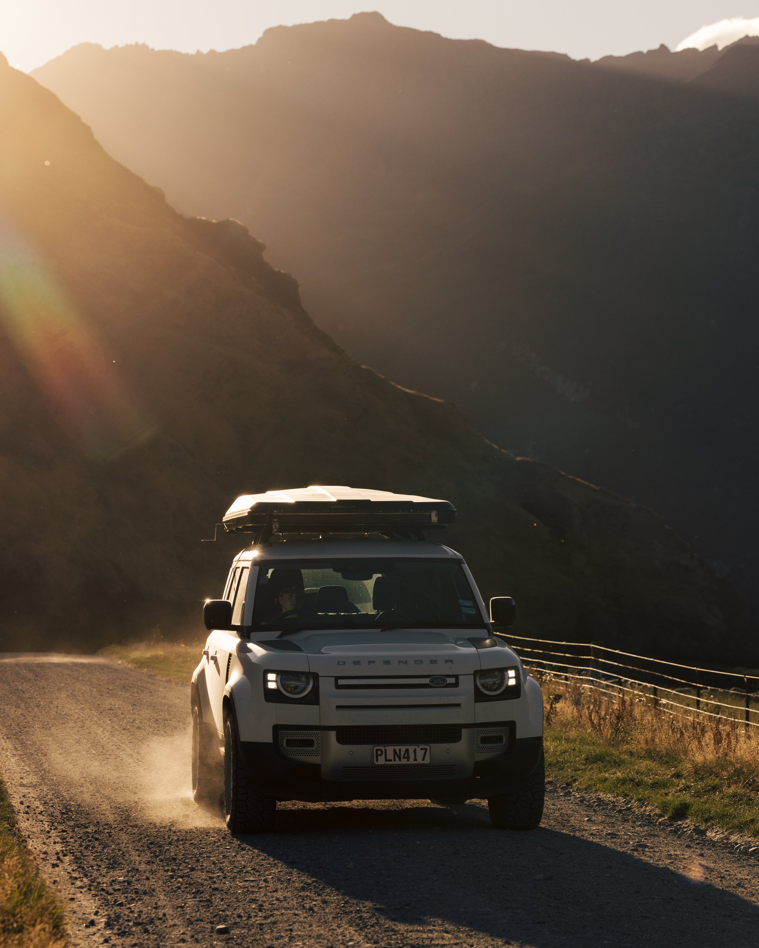 A white Land Rover Defender driving on a dirt road in a mountainous landscape during sunset.