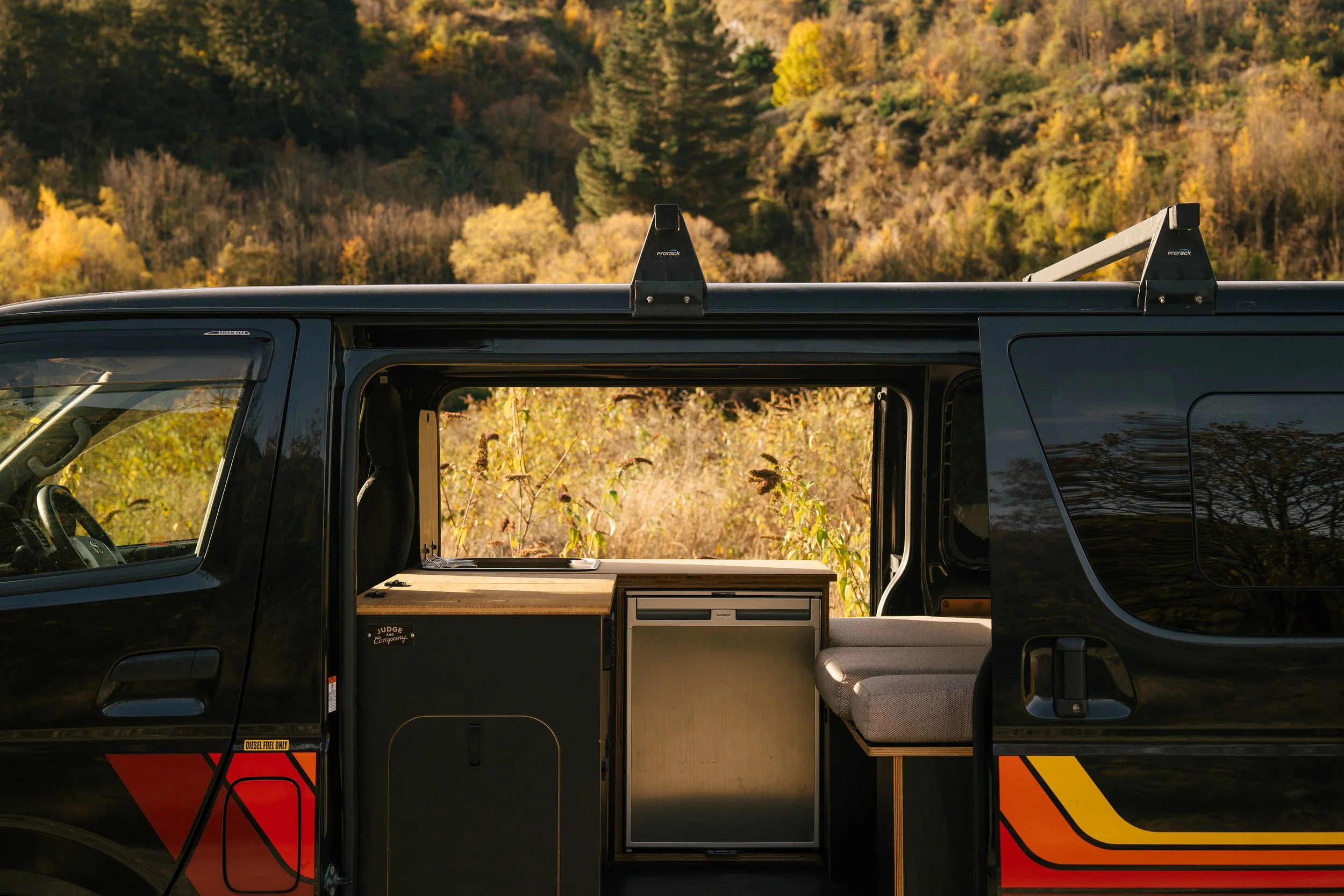 The side view of a black camper van with a mountain landscape and trees with fall foliage in the background. The van's sliding door is open, revealing an interior with a small kitchen area and seating.