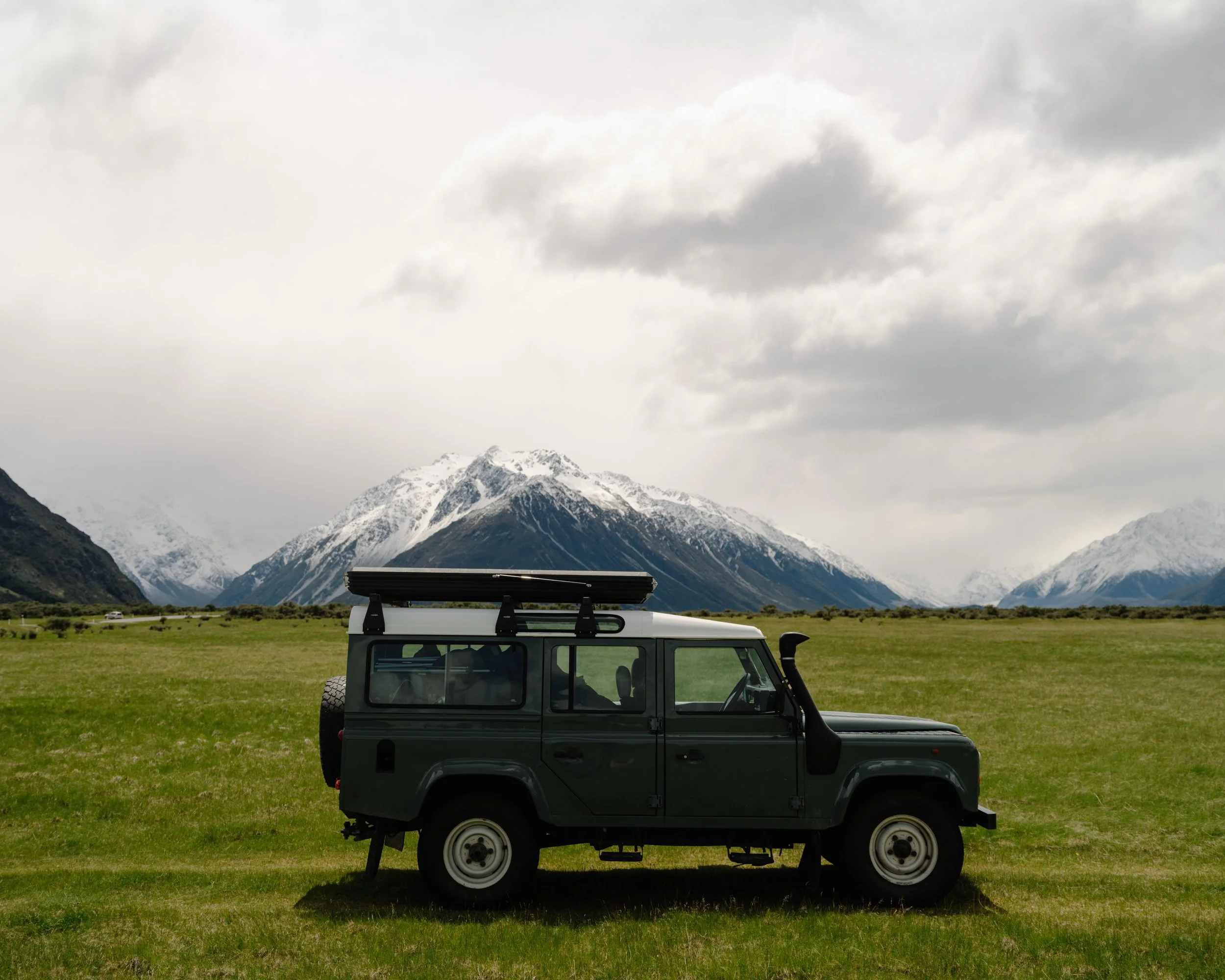 A black off-road vehicle parked on a grassy field with snow-capped mountains in the background and cloudy sky overhead.
